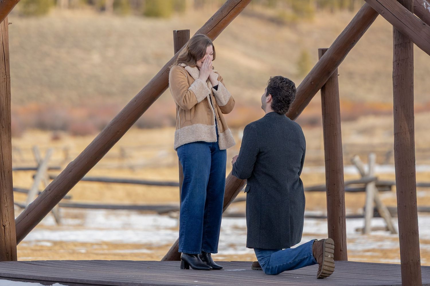 Man proposes on one knee to woman on wooden bridge in scenic winter landscape.