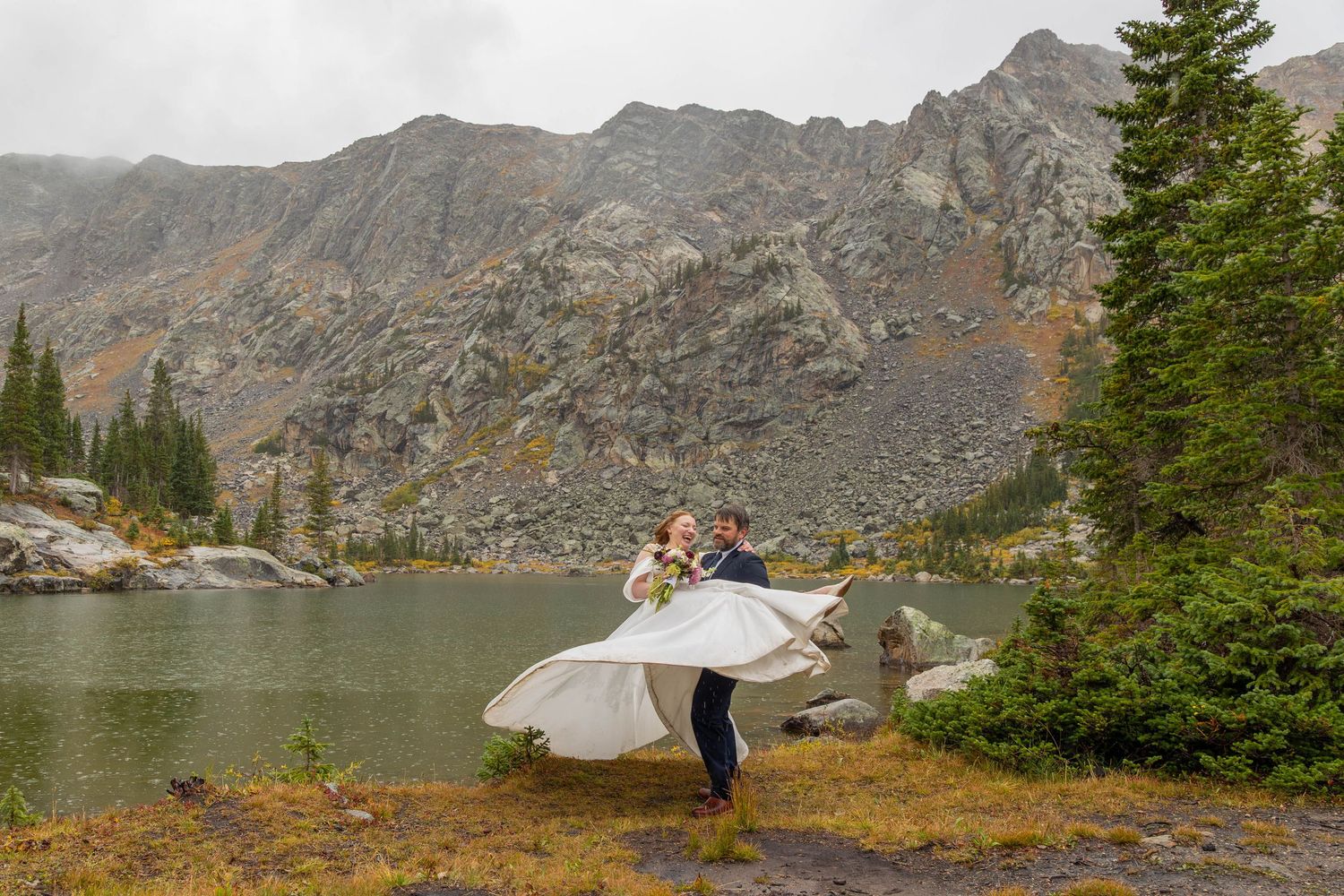 Bride in a flowing white gown twirls with her groom beside a calm alpine lake, surrounded by evergreen trees and rugged mountain cliffs.