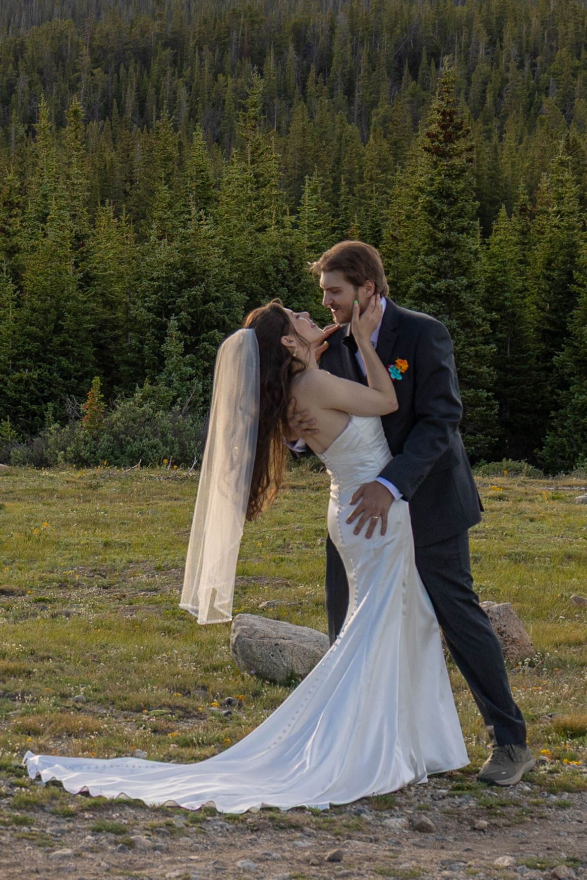 Bride and groom share romantic kiss outdoors with evergreen forest backdrop during mountain wedding ceremony.