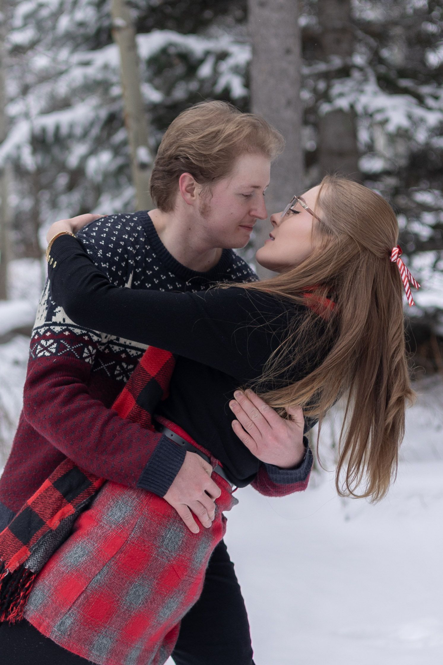 Couple sharing a romantic moment in a snowy winter forest, wearing cozy holiday sweaters and embracing tenderly.