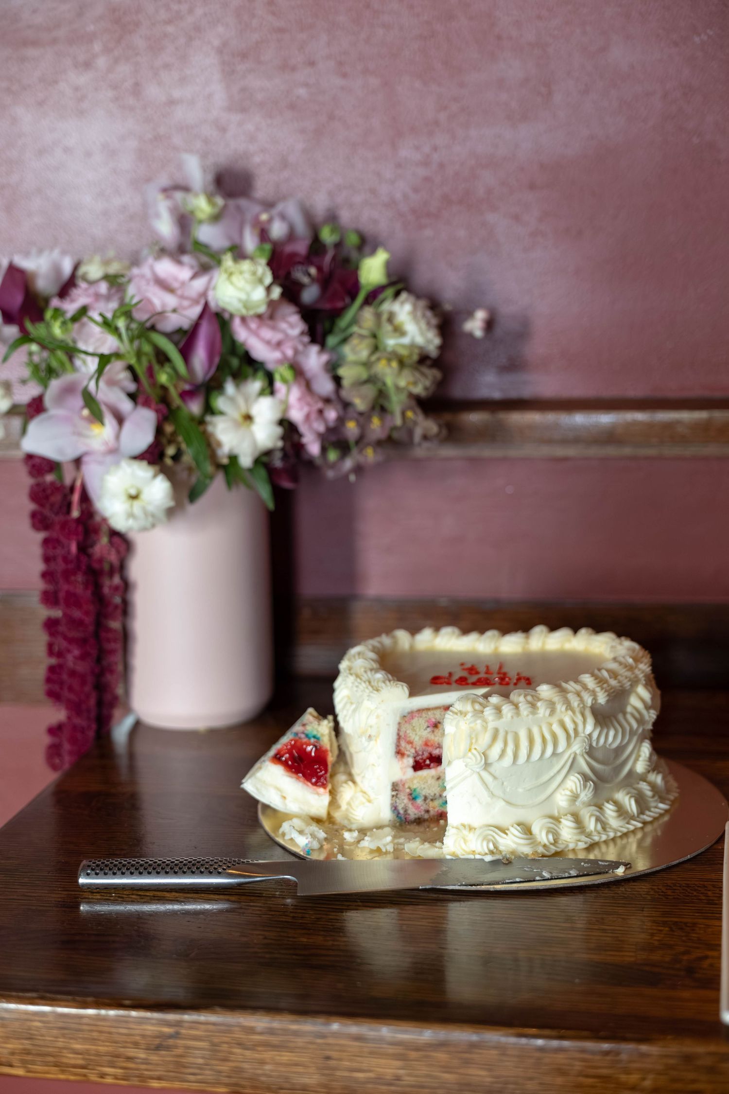 A sliced heart-shaped cake with white frosting and berry filling on a wooden table beside a pink vase of flowers.