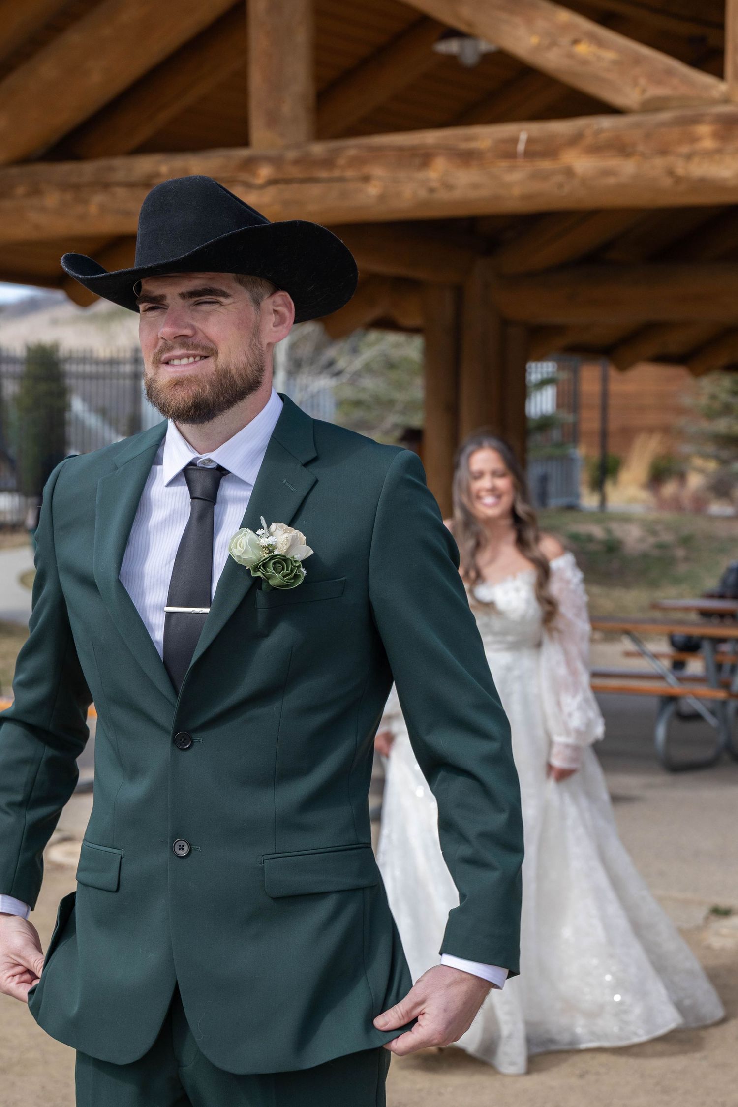 Groom in green suit and black cowboy hat smiles as bride in white lace gown approaches during outdoor wedding ceremony.