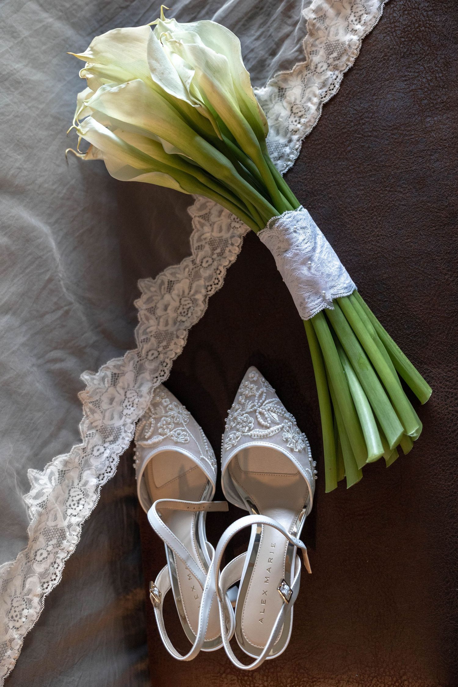 White lace bridal shoes and calla lily bouquet tied with ribbon displayed on brown leather surface with lace veil.