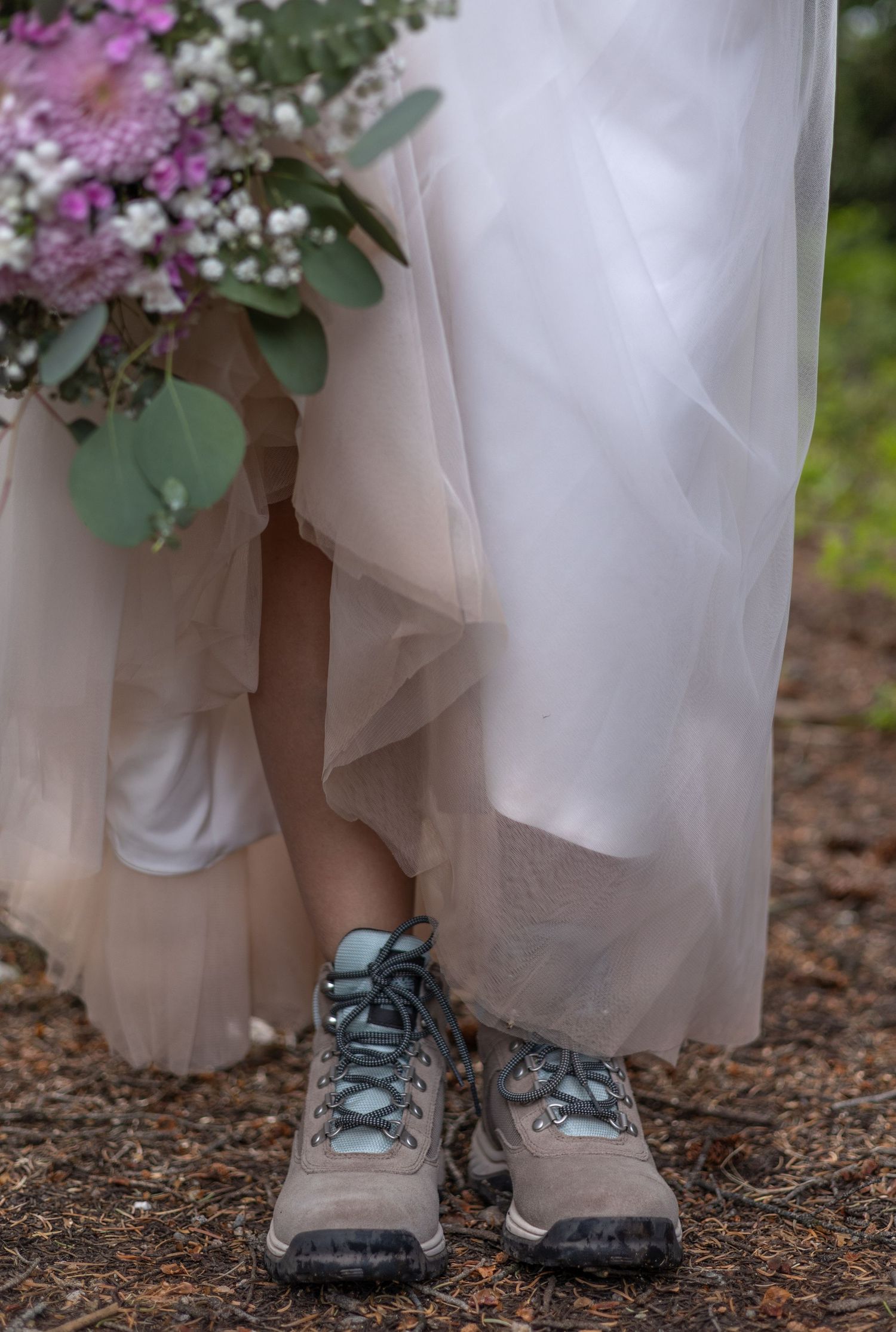 A bride wearing hiking boots under her white wedding dress stands on a dirt path in a playful outdoor photo series.