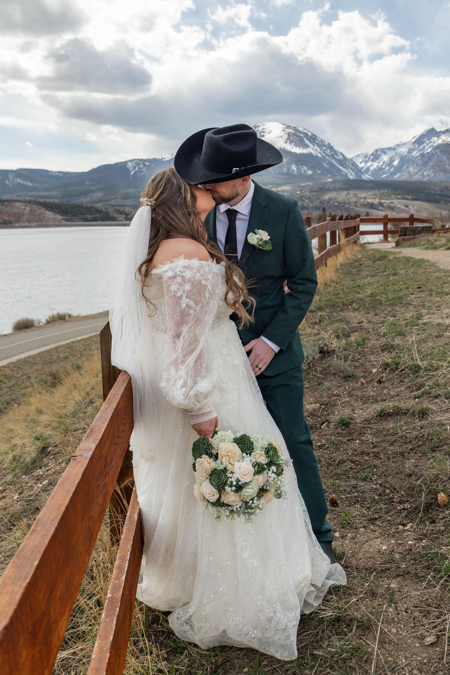 Bride and groom kissing outdoors near a mountain lake, she holds white bouquet, he wears green suit and black cowboy hat.