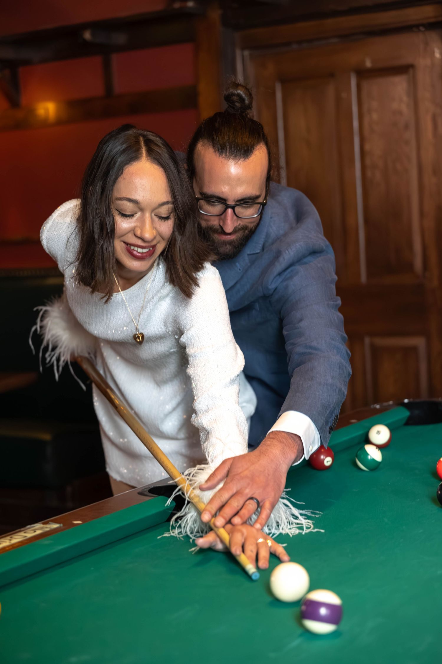 Couple playing pool together at a bar, man guiding woman's billiard shot over green felt table.