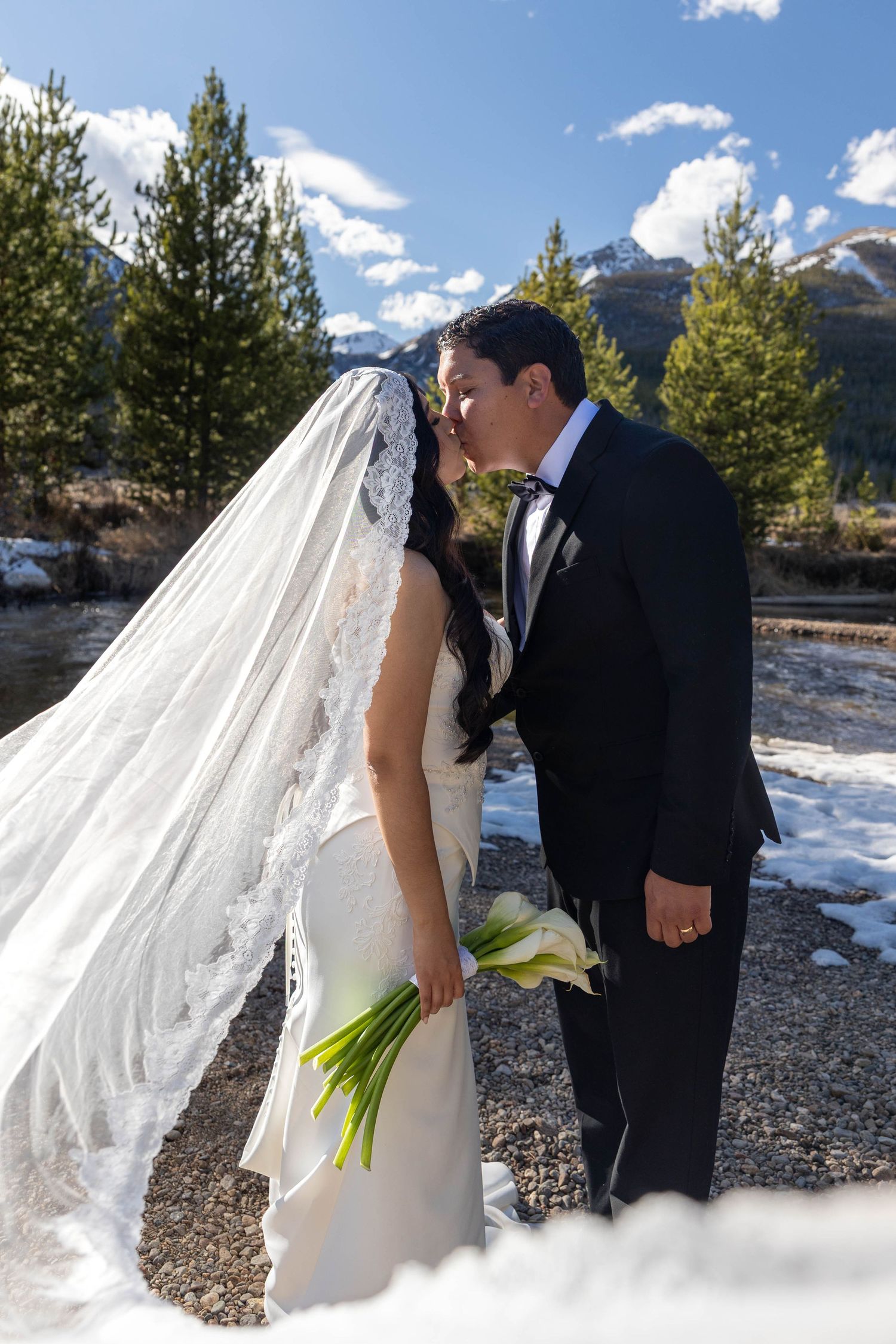 Bride and groom share a kiss outdoors in a snowy mountain setting, bride holding white calla lilies with veil flowing.