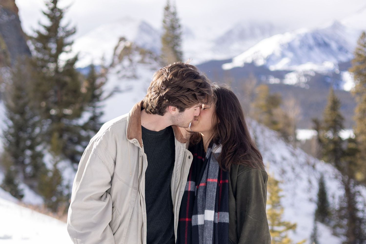 Couple sharing a romantic kiss in a snowy mountain landscape, wearing winter coats and a plaid scarf.