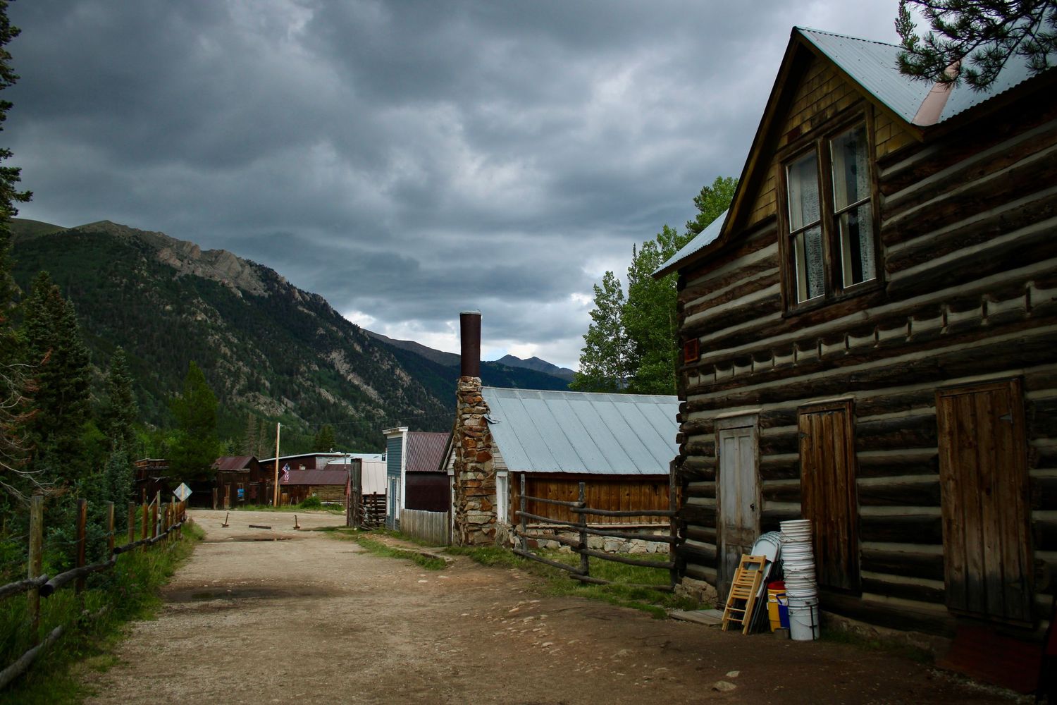 Rustic log cabin buildings along a dirt road with dramatic storm clouds over mountain peaks.