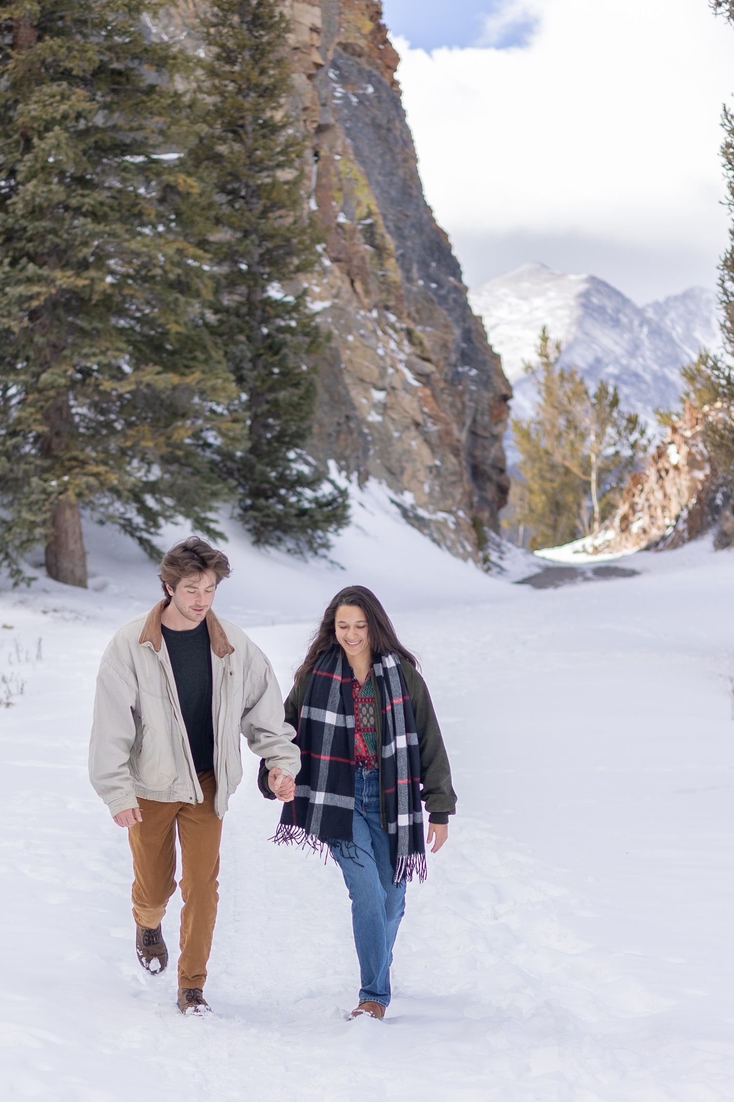 Couple holding hands walking through snowy mountain landscape with pine trees and rocky peaks in winter.