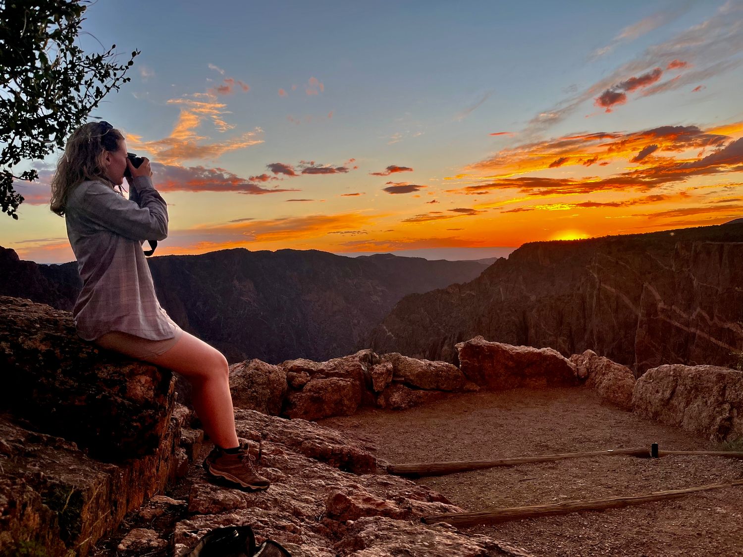 A silhouetted figure sits on a cliff edge photographing a dramatic orange and purple sunset over a canyon landscape.