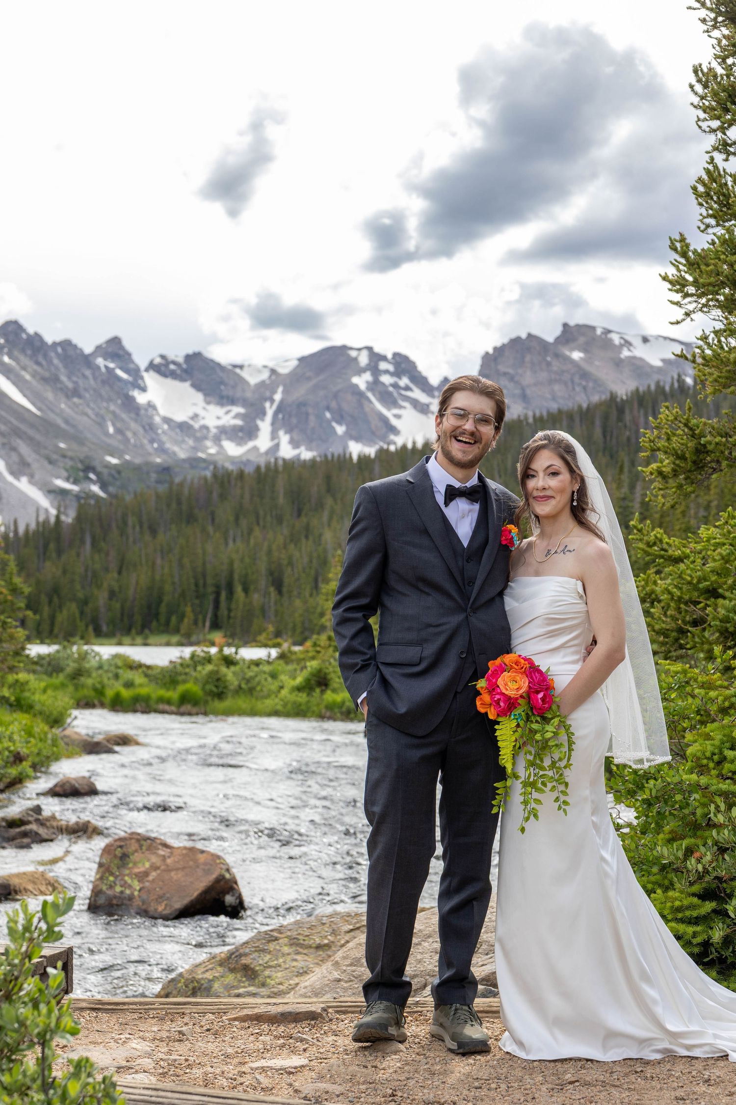 Bride and groom posing by a mountain stream with snow-capped peaks and evergreen forest backdrop on their wedding day.