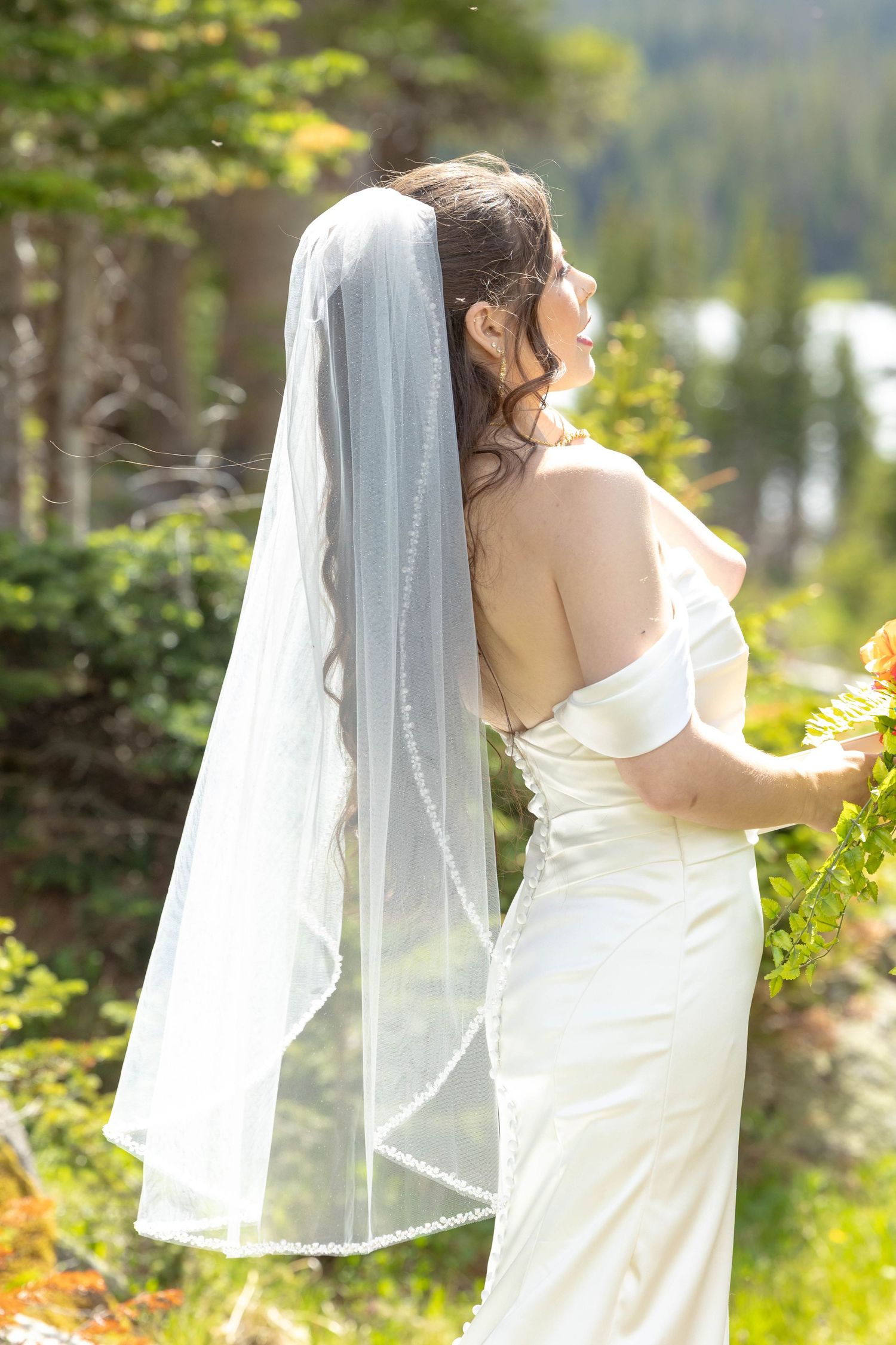 Bride in white off-shoulder gown and pearl-trimmed veil stands outdoors among green trees, holding a bouquet.