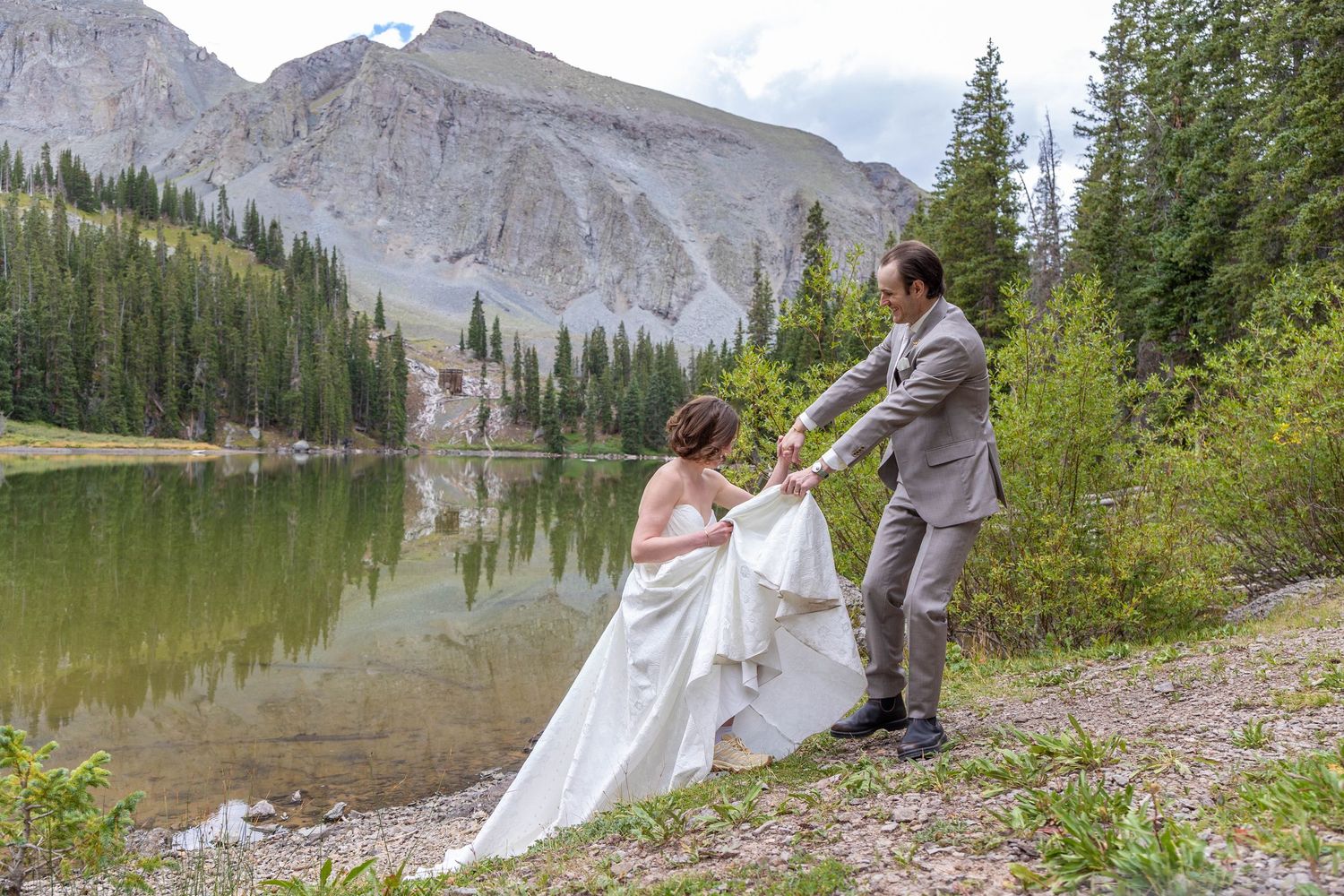 Bride in a flowing white gown and groom in a gray suit hold hands beside a calm mountain lake with pine trees and rocky peaks.