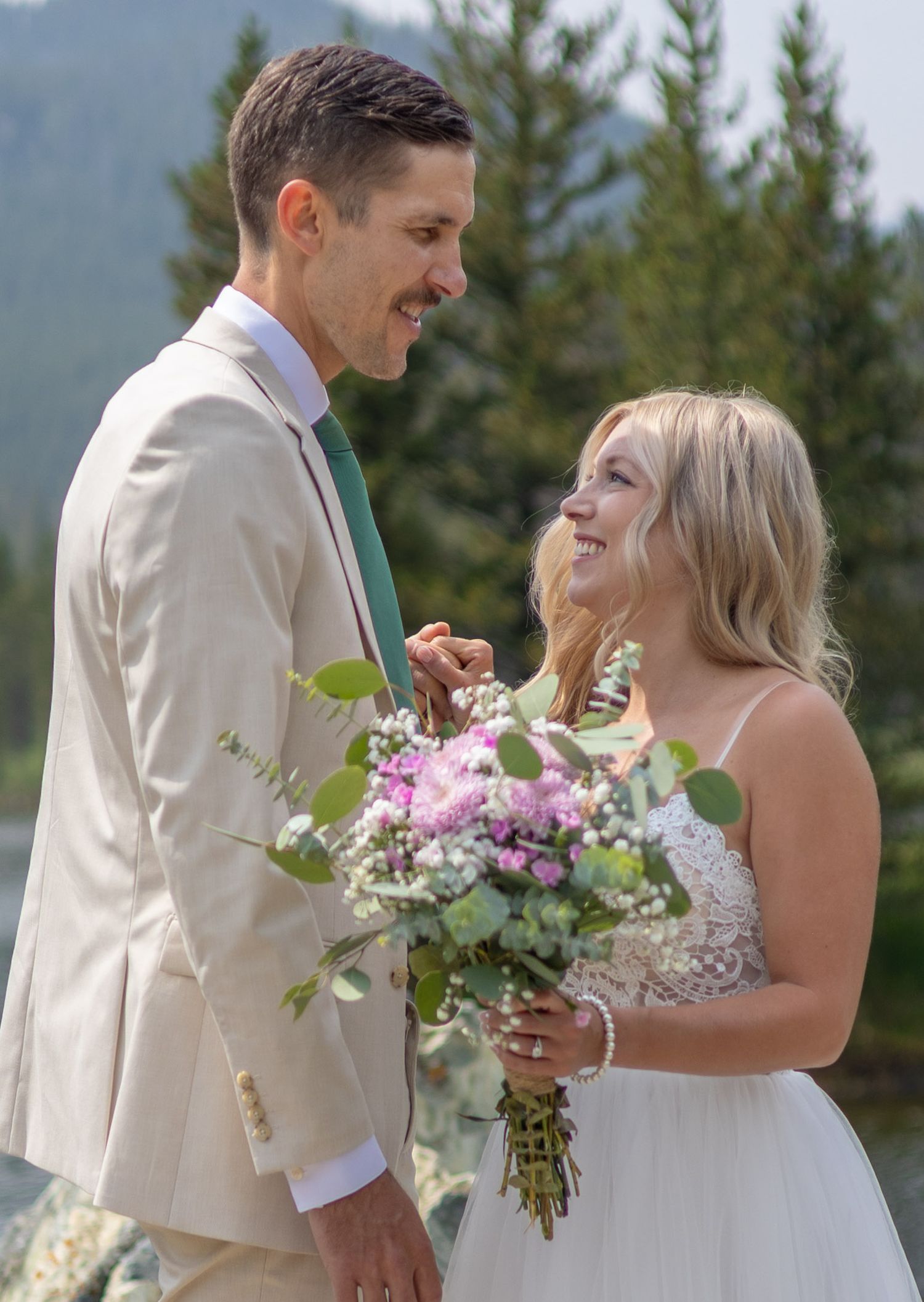 A bride holding a floral bouquet and groom in a tan suit share a joyful moment outdoors with pine trees behind them.