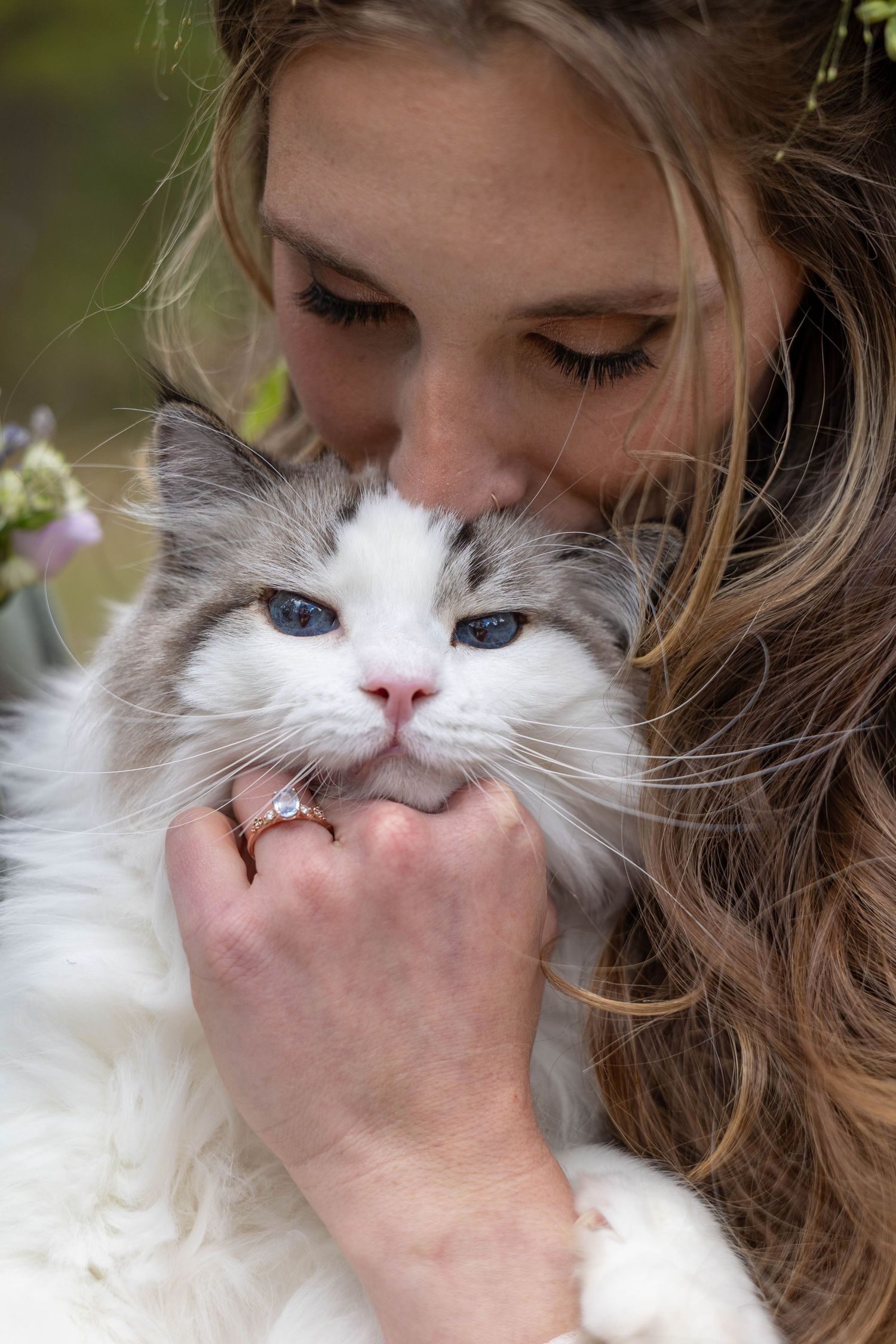 Woman with long hair kissing a fluffy white cat with blue eyes while wearing a diamond engagement ring.