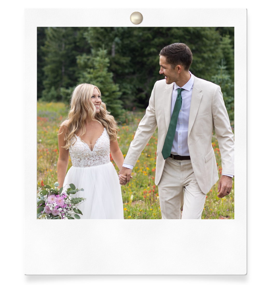 A smiling bride in a white gown holds purple flowers while walking hand-in-hand with a groom in a tan suit in a meadow.