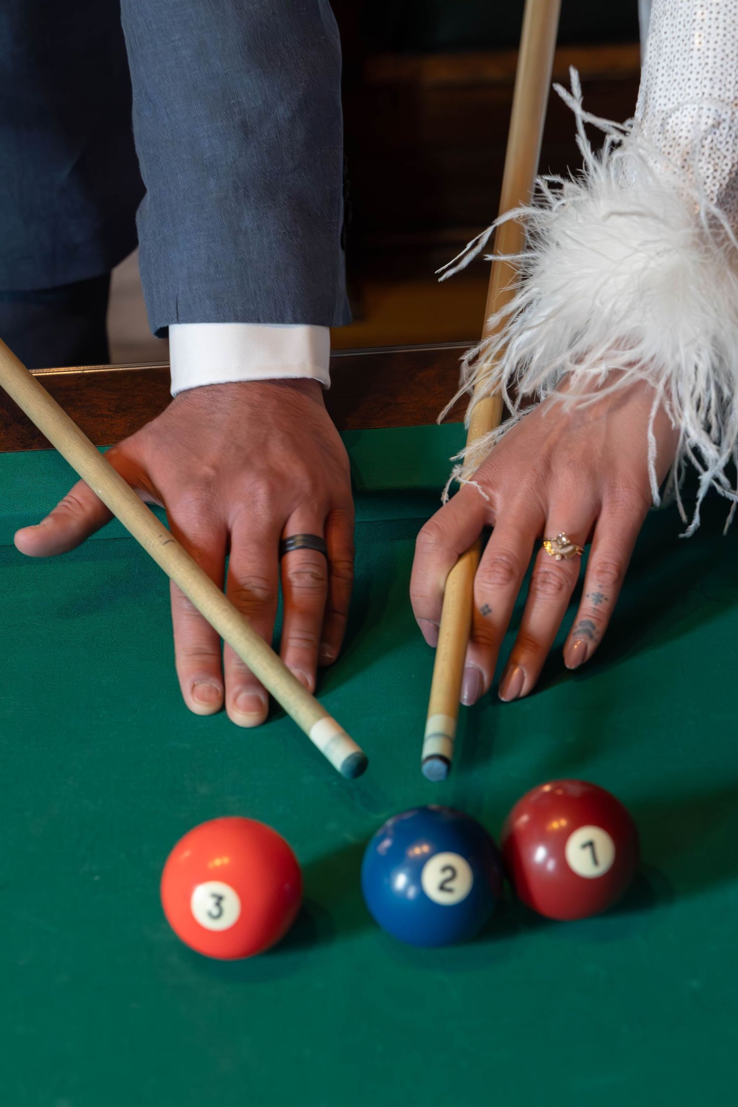 Newlywed couple playing billiards, hands with wedding rings holding cues near balls numbered 1, 2, and 3 on green table.