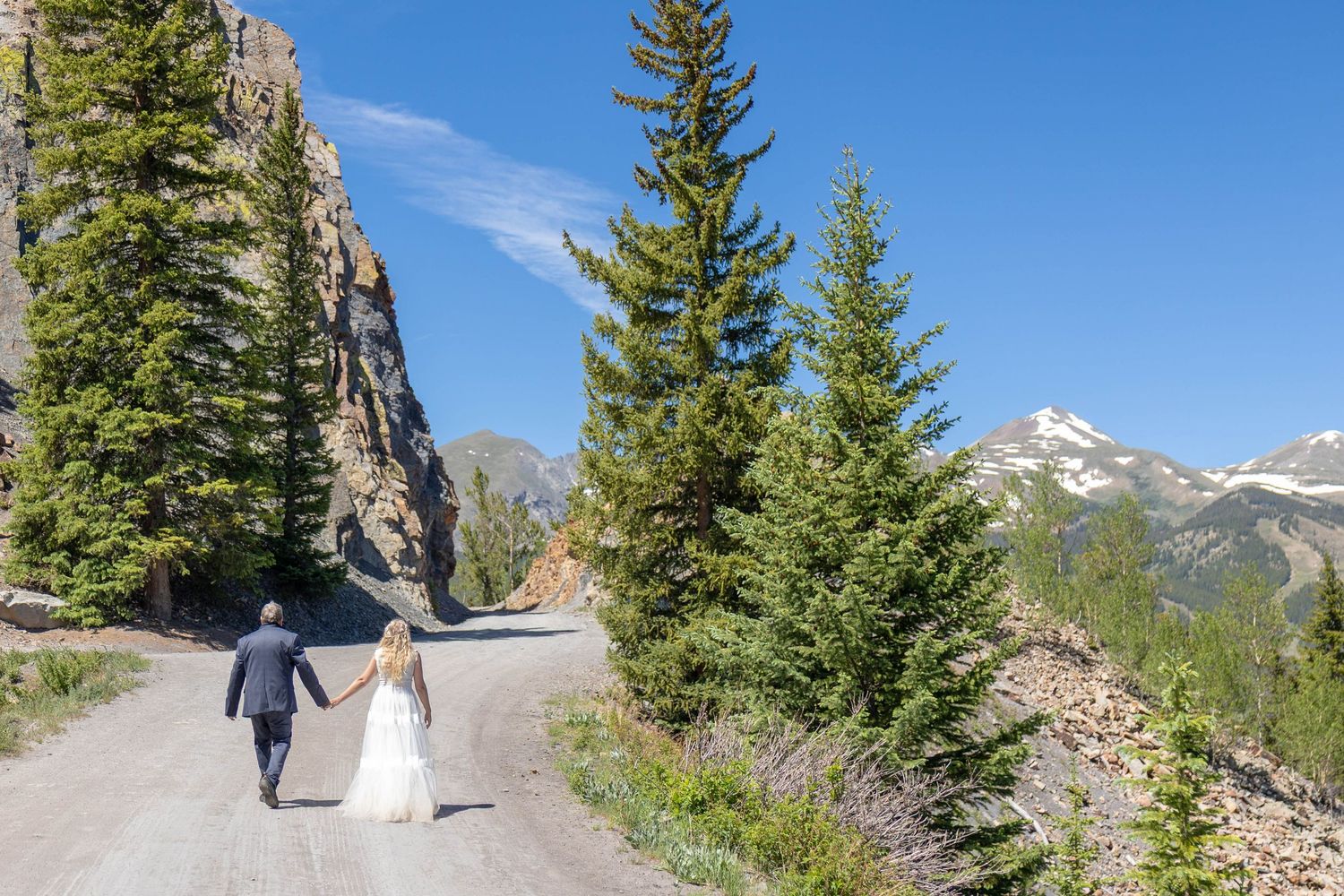 Bride and groom walk hand-in-hand on a mountain road surrounded by pine trees and rocky peaks under a clear blue sky.