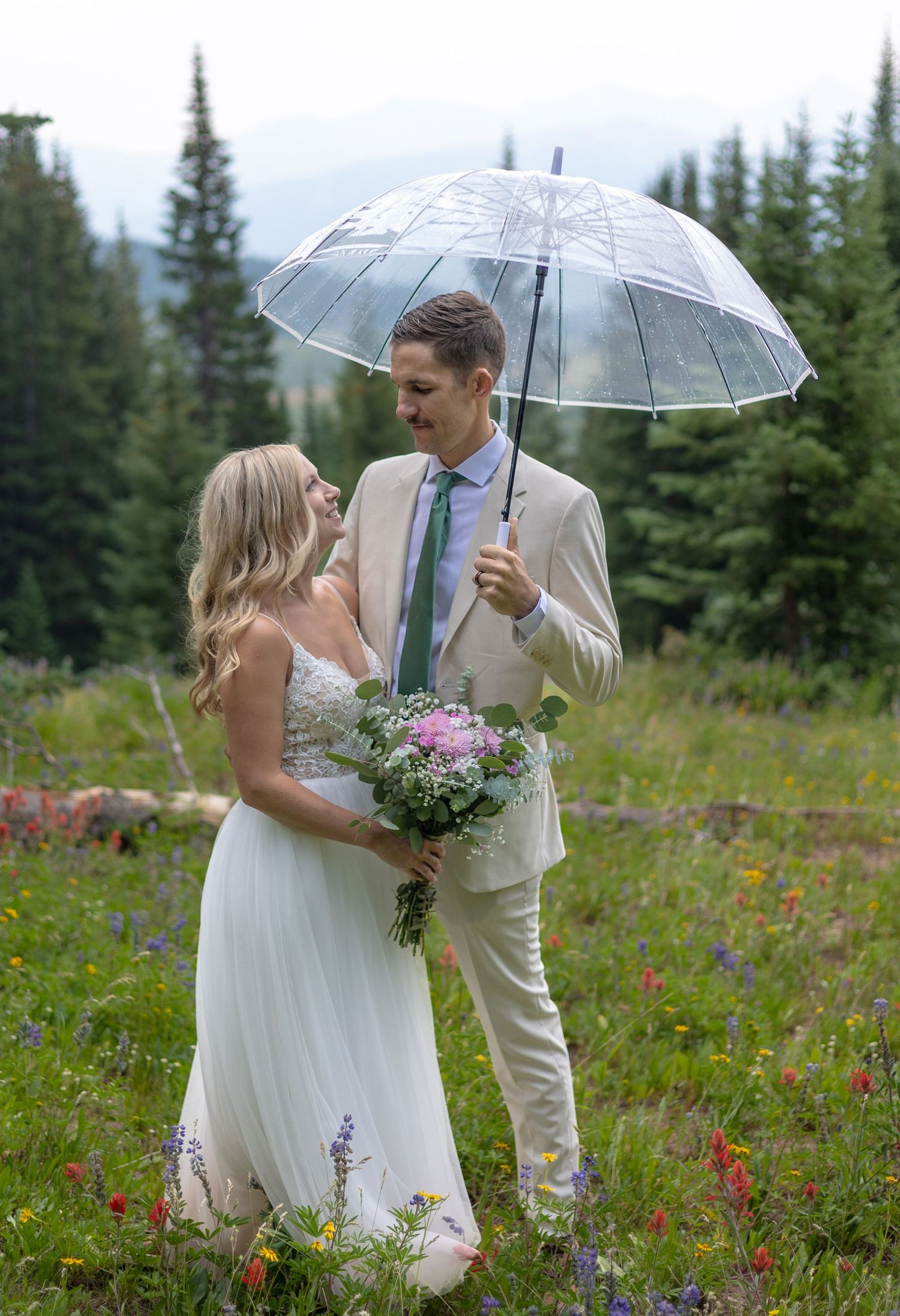 Bride and groom share a tender moment under a clear umbrella in a wildflower meadow with pine trees backdrop.
