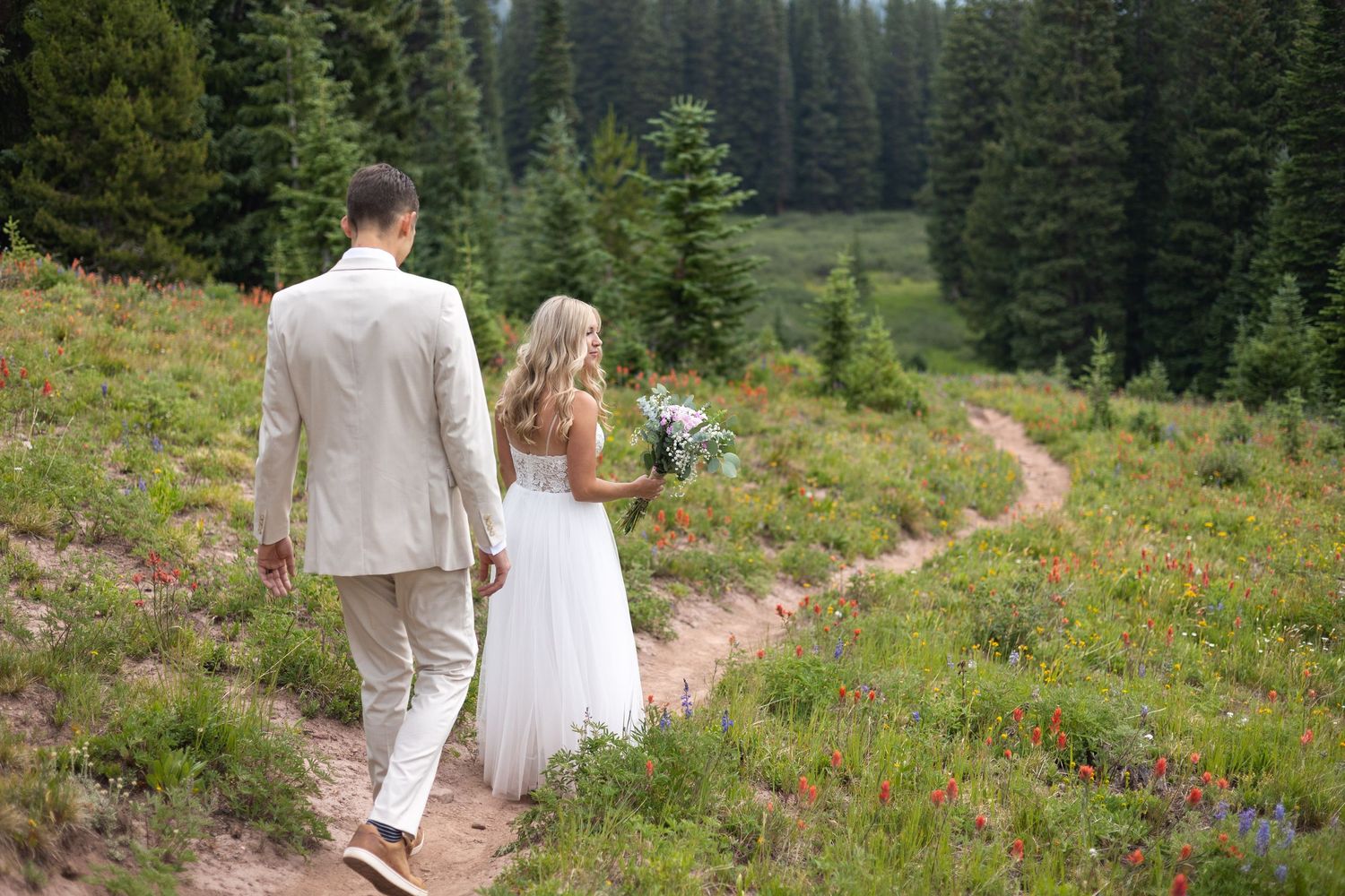 Bride and groom walking hand in hand along a mountain trail surrounded by wildflowers and evergreen trees.