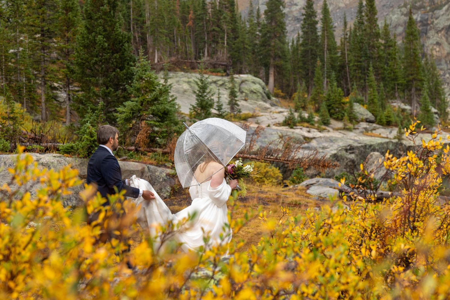 Bride and groom walk through golden autumn foliage in Rocky Mountain forest, bride holding clear umbrella on rainy wedding day.