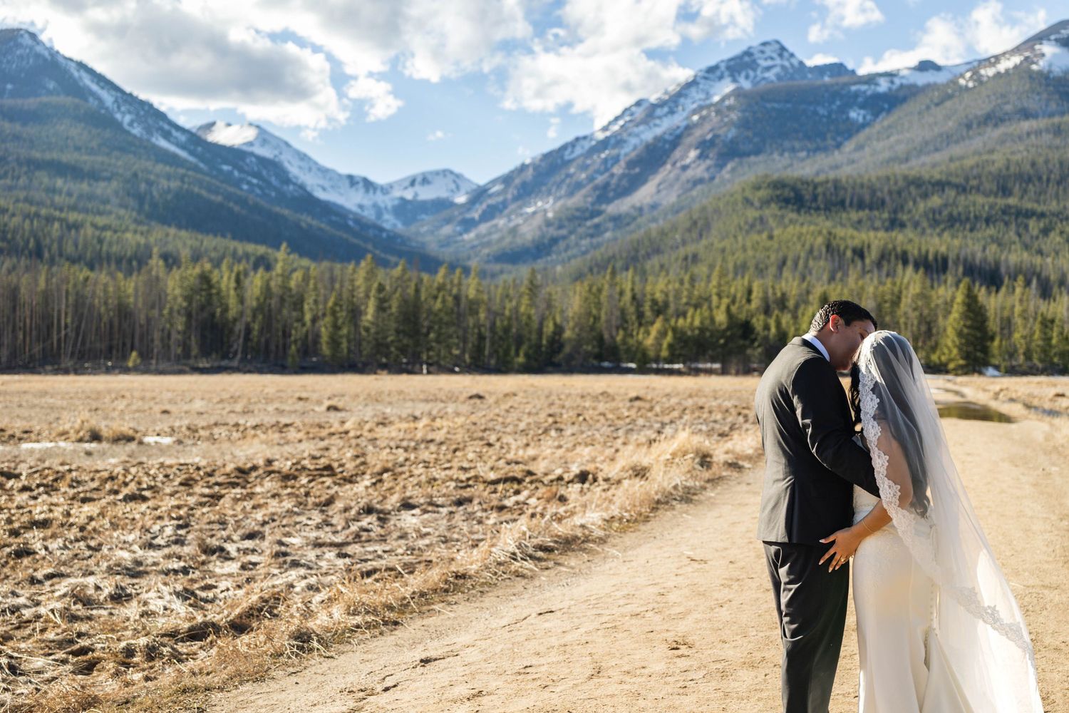 A bride and groom share a romantic kiss with stunning Rocky Mountain peaks and evergreen forests as their backdrop.