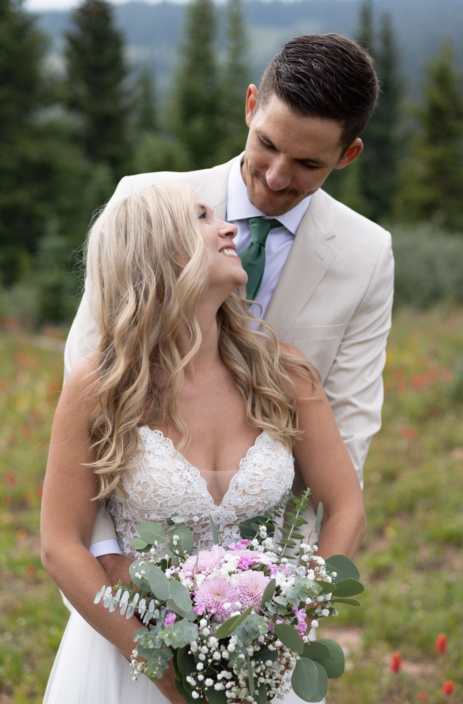 A couple in wedding attire shares romantic moments on a mountainside with evergreen trees and wildflowers in the background.