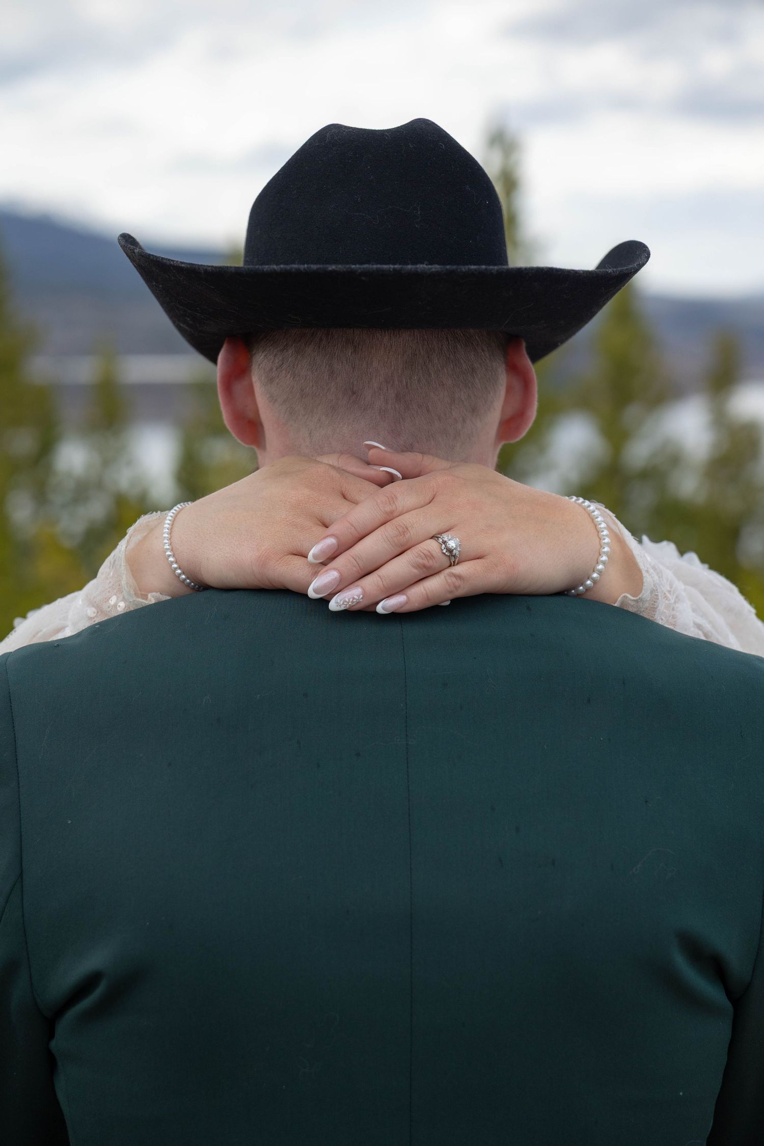 Bride places her hand on groom's neck as they embrace, groom wearing black cowboy hat and green suit at outdoor wedding.