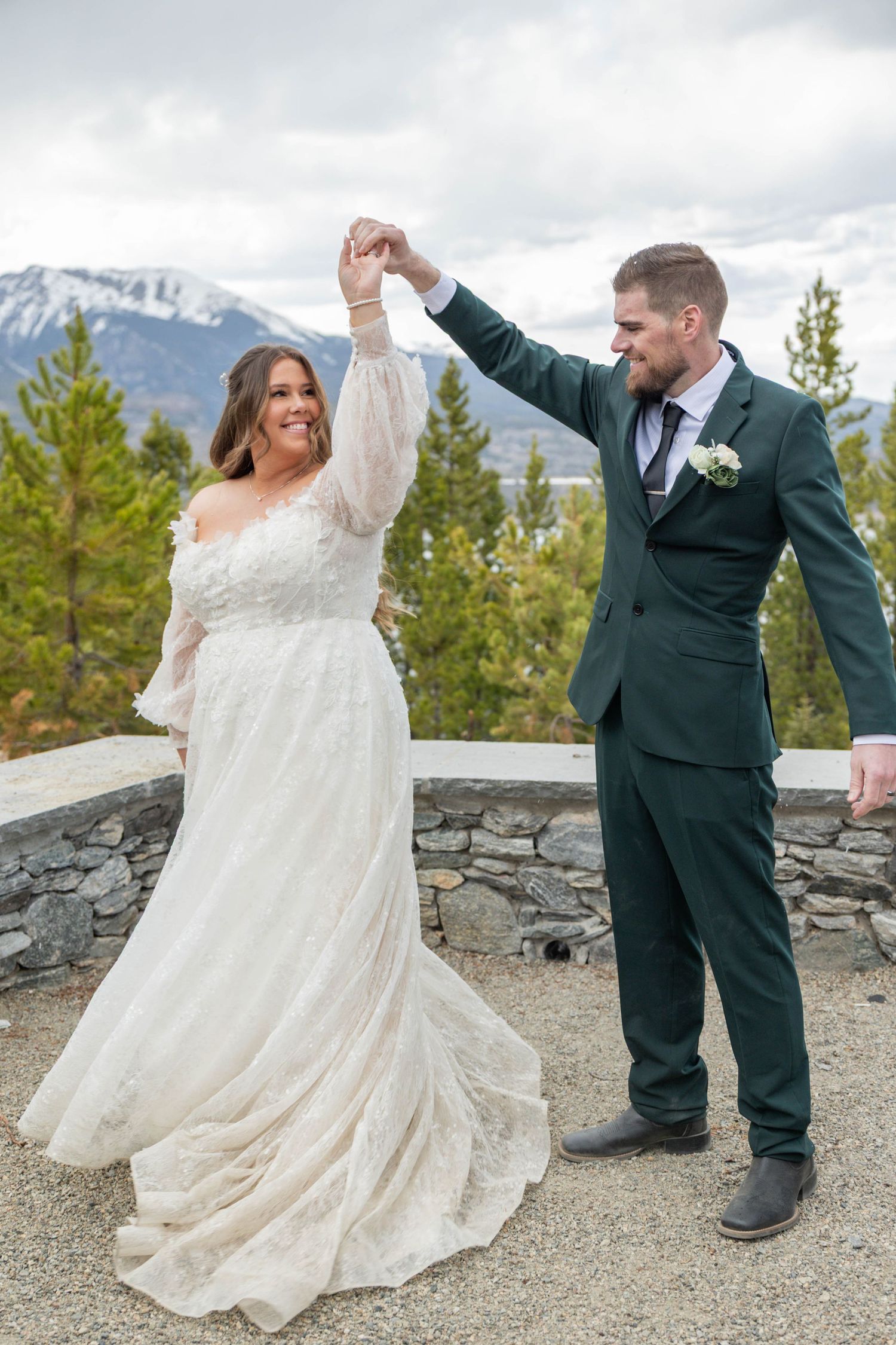 A joyful bride and groom dance outdoors with mountain and forest views, she in lace gown, he in dark green suit.