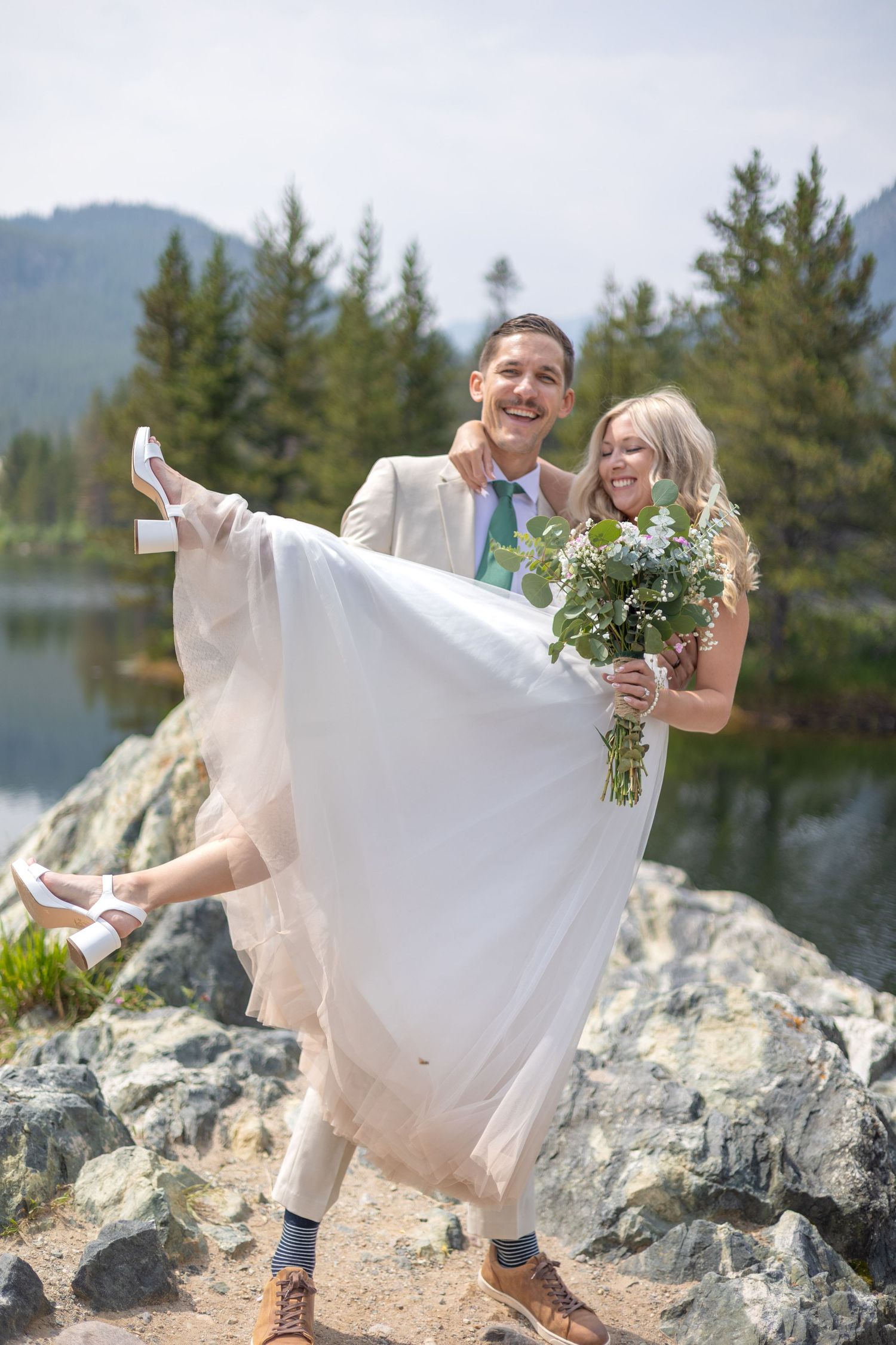 A couple in wedding attire playfully poses on rocky terrain by a lake with evergreen trees in the background.