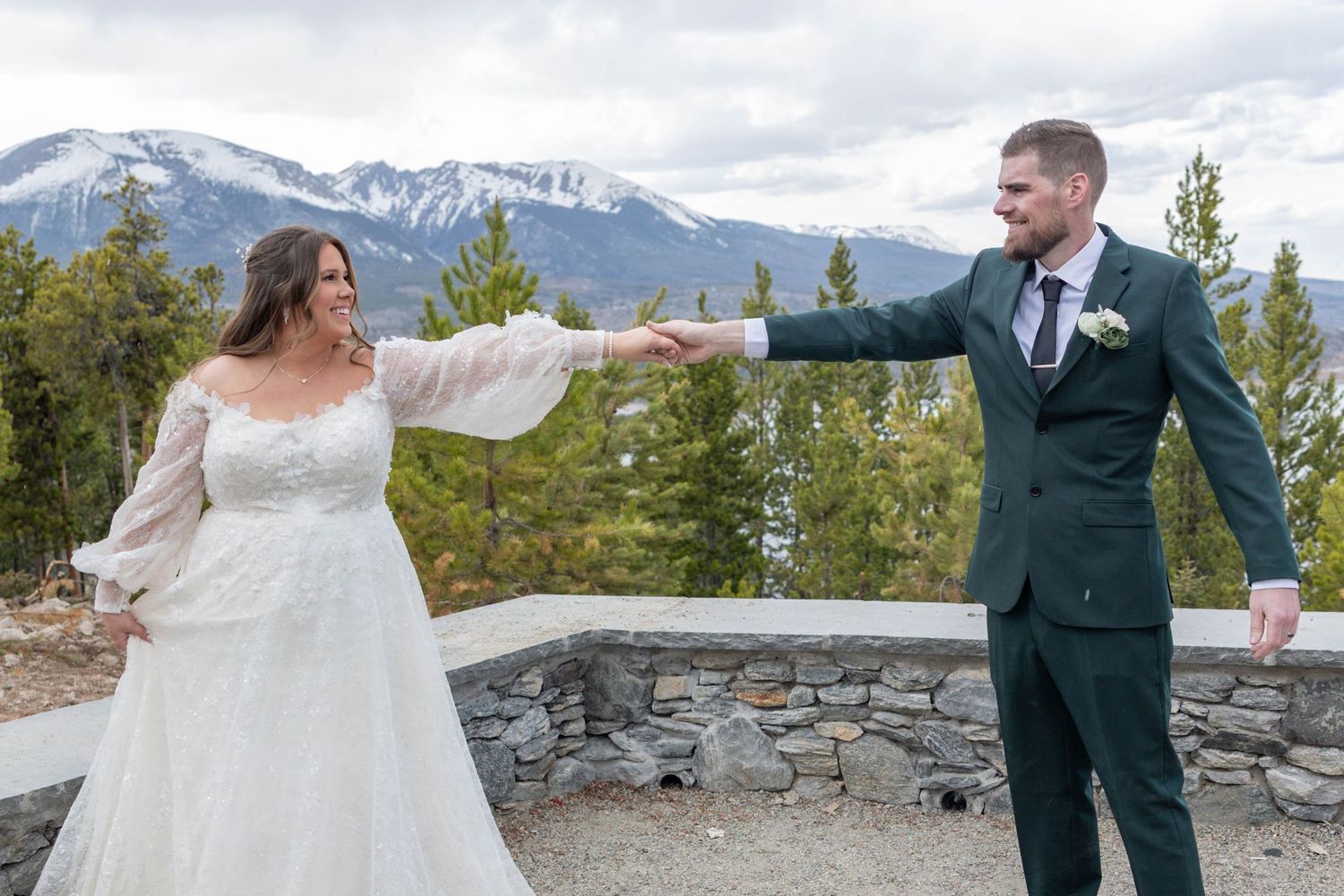Bride and groom hold hands outdoors with snow-capped Rocky Mountains backdrop at a scenic mountain wedding venue.