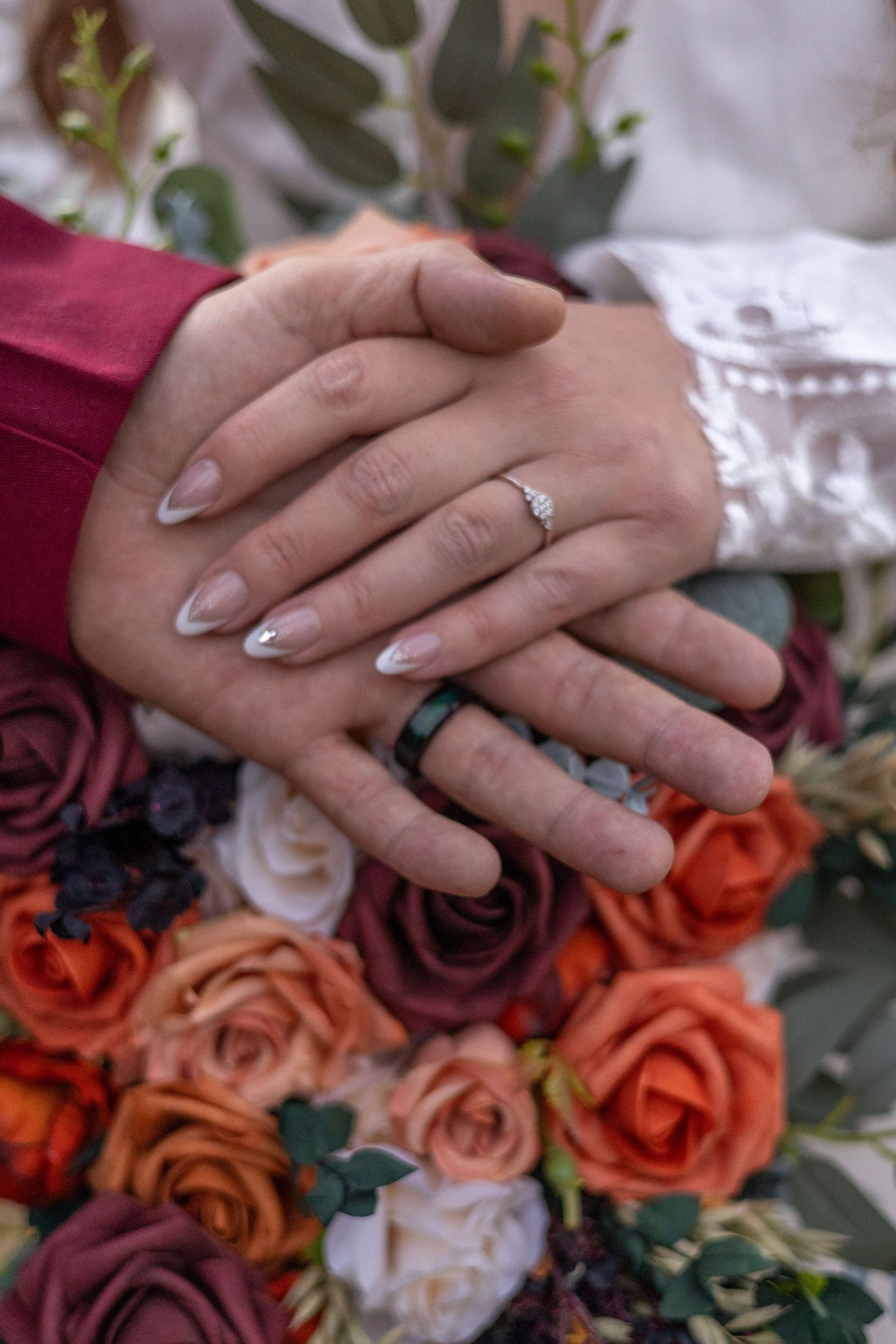 Close up of wedding rings and hands resting on orange and burgundy rose bouquet.