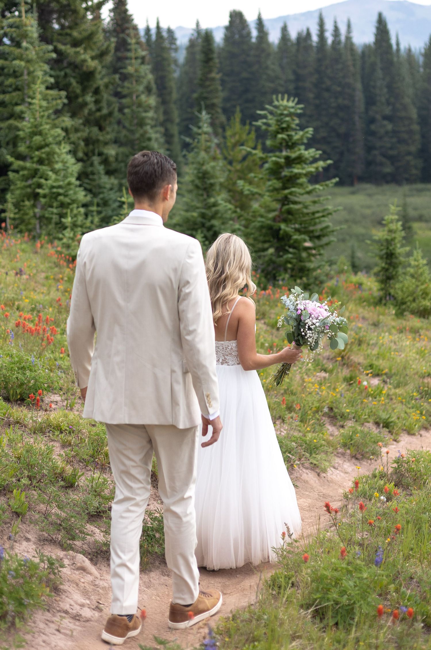 A couple in wedding attire walk along a mountain trail surrounded by evergreen trees and wildflowers in Colorado.