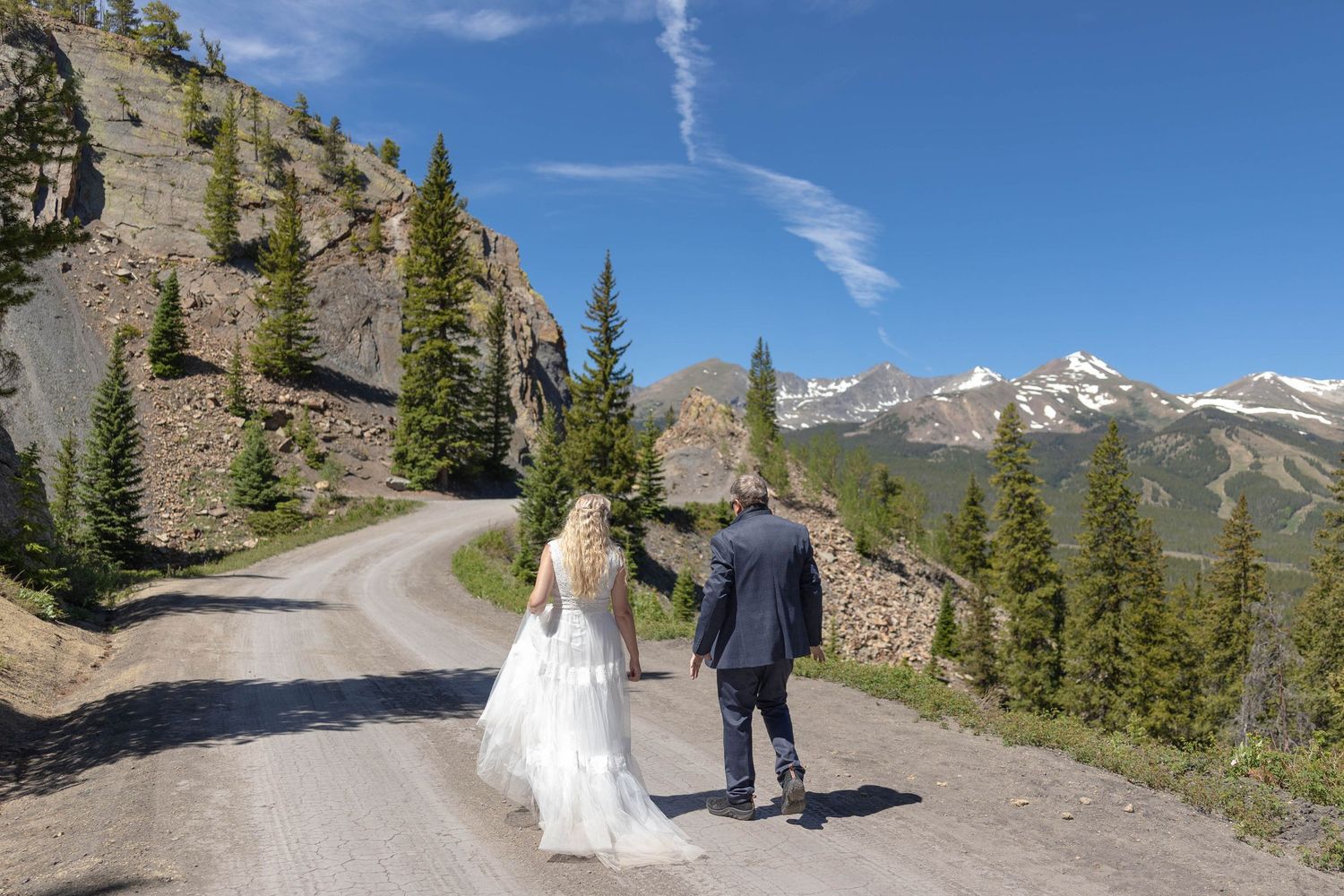 Bride in a flowing white wedding dress and groom in a dark suit walk a winding mountain dirt road with pine trees and snowy peaks behind.