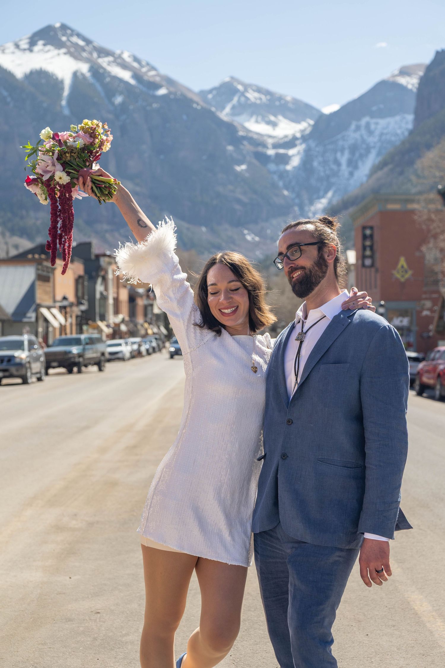 Joyful newlyweds pose on a mountain town street, bride in white mini dress holding bouquet aloft, groom in blue suit.