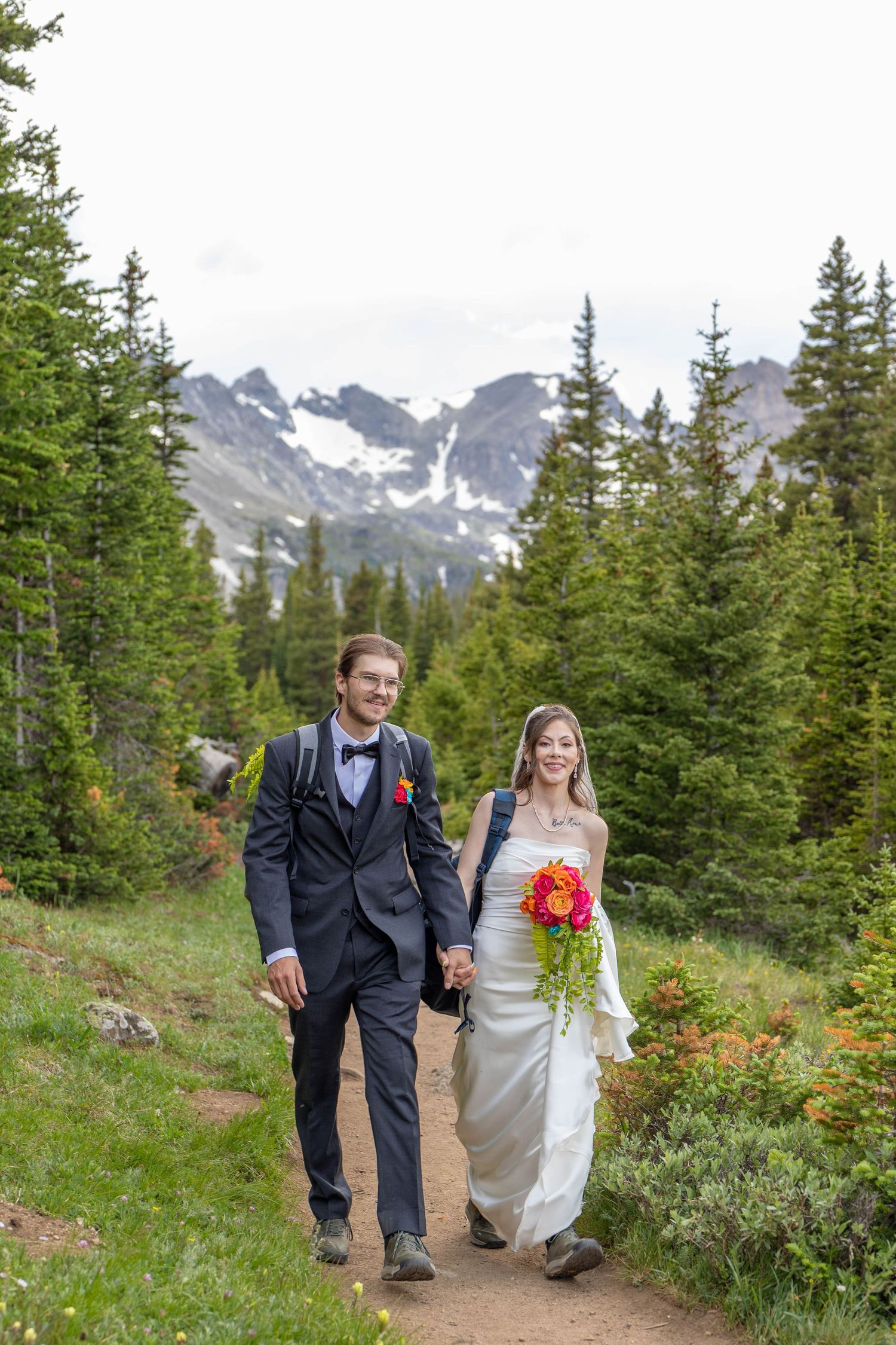 Bride and groom holding hands walking on mountain trail in wedding attire with bouquet and snowy peaks backdrop.