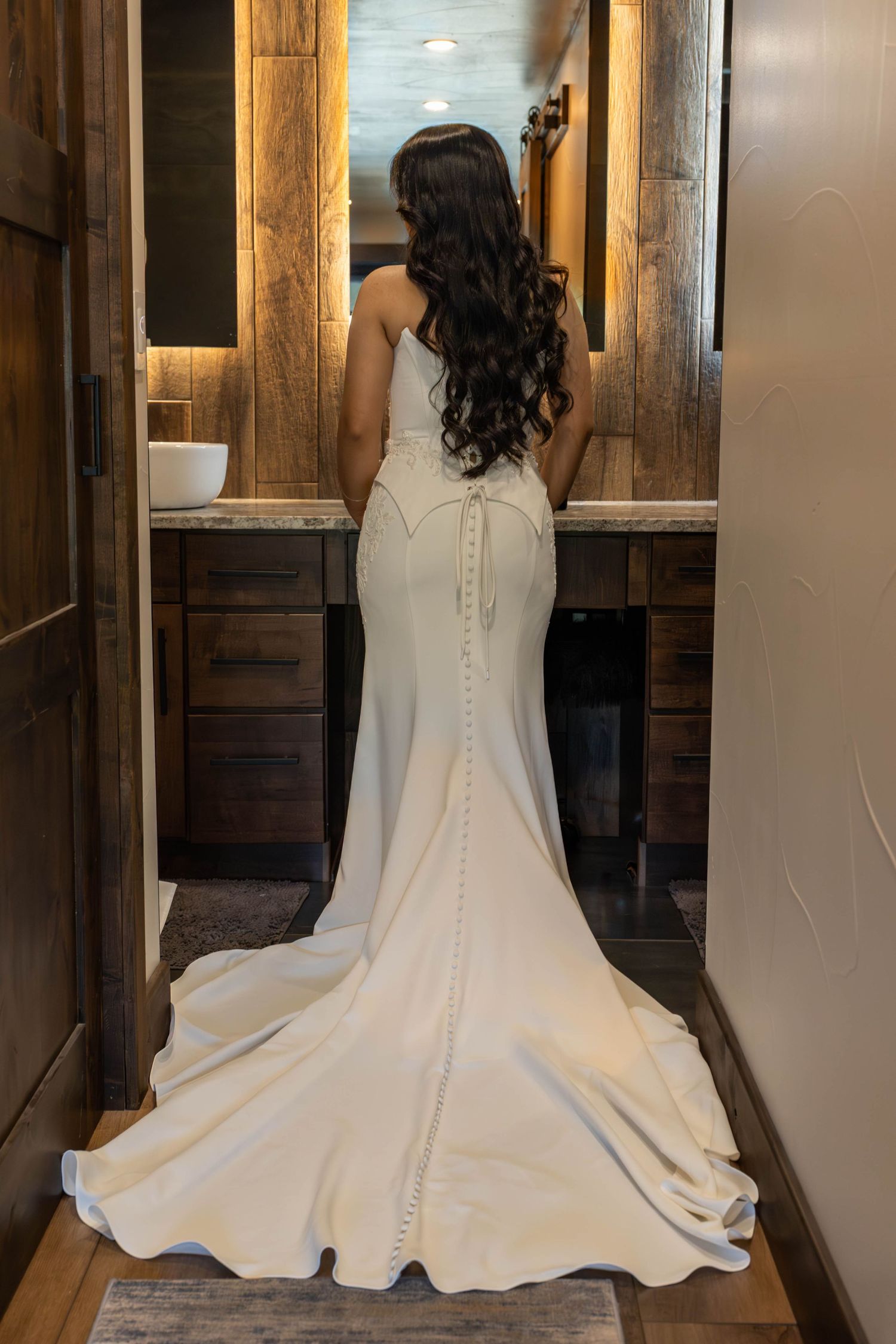 Bride in elegant white mermaid wedding gown with button-back detail and train, viewed from behind in a rustic bathroom.