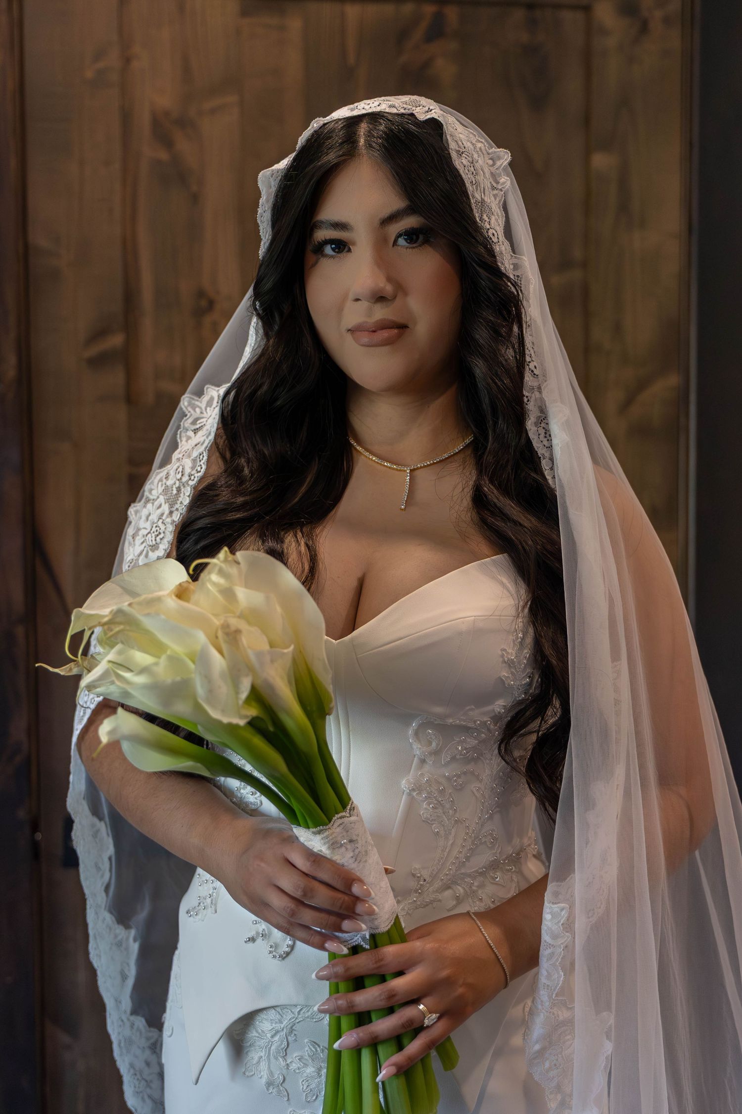 Bride in white strapless gown and lace veil holding white calla lily bouquet with gold necklace against wood backdrop.