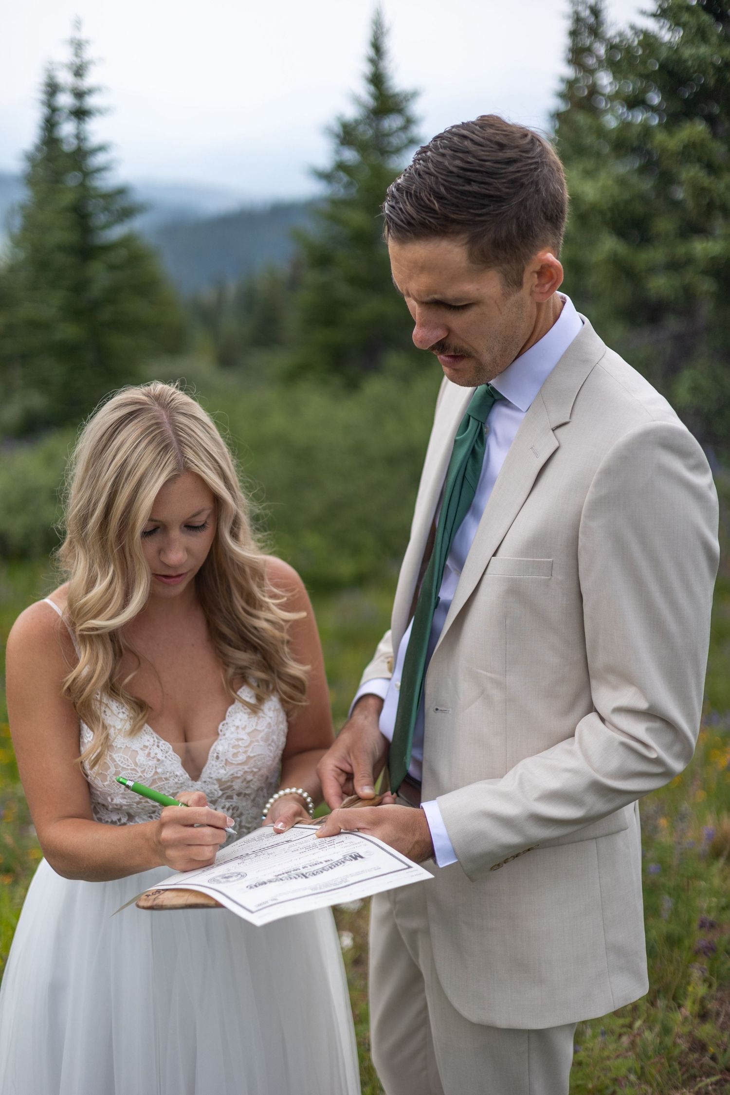 A couple in wedding attire signs documents together on a mountaintop surrounded by evergreen trees.