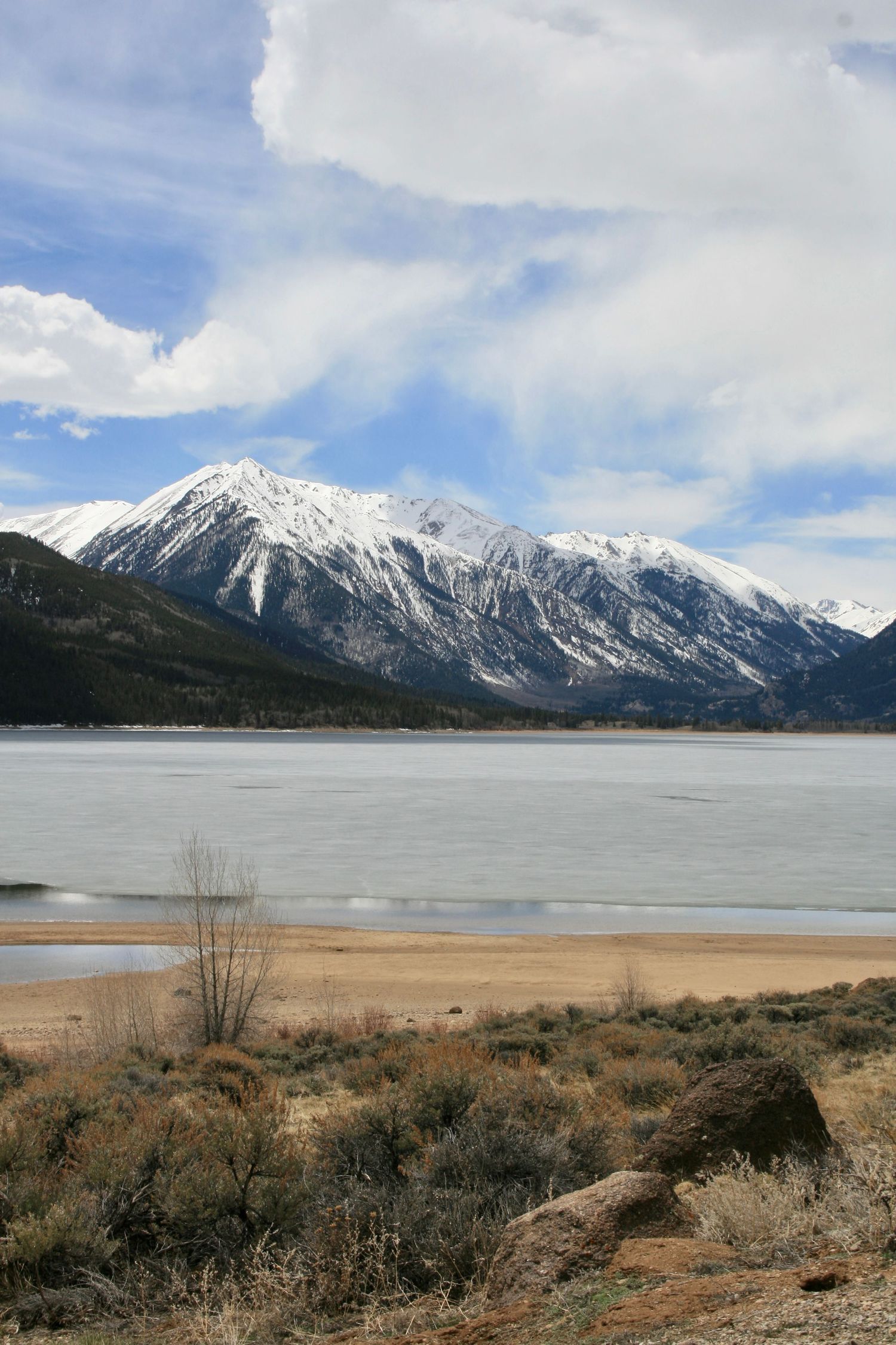 Snow-capped mountain peaks tower over a frozen lake with sandy shoreline and sage brush in the foreground. Twin Lakes, Leadville