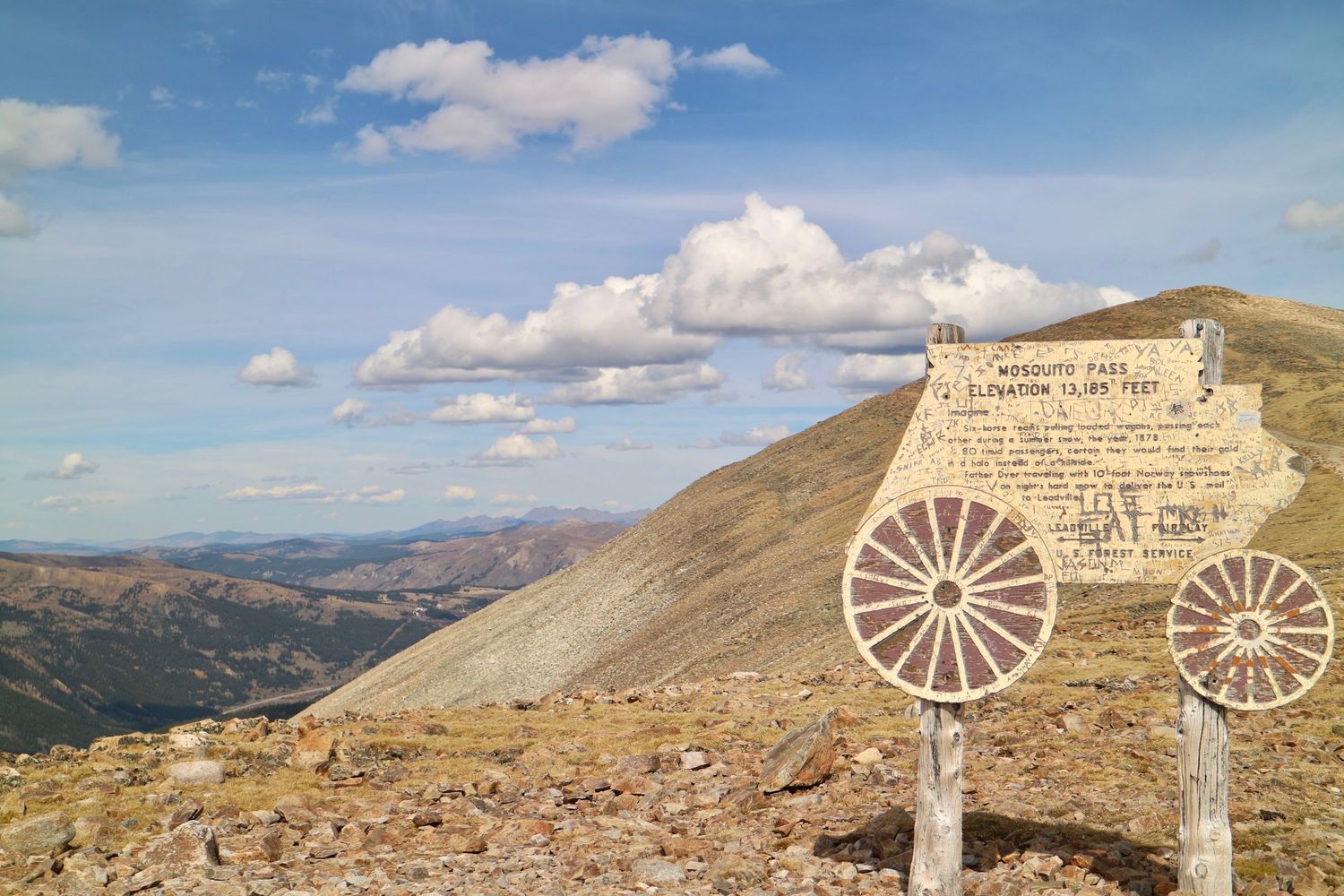 Wooden mountain pass sign with wagon wheel decorations on a rocky alpine summit overlooking rolling ridges under a blue sky with clouds.