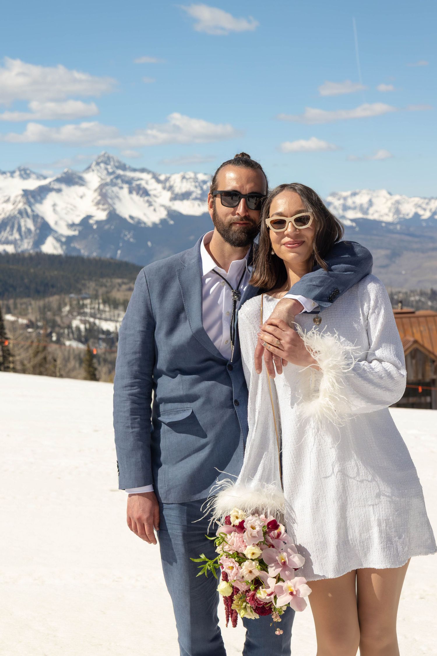 Couple in wedding attire poses in snowy mountain landscape, bride in white mini dress holding floral bouquet.