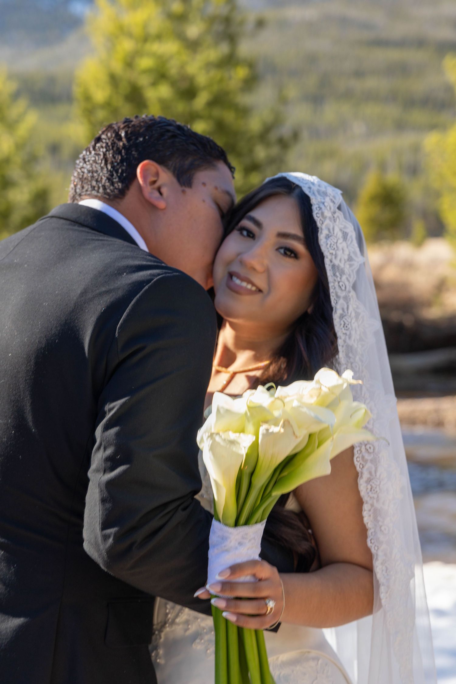 Groom in black suit kissing bride on cheek as she smiles holding white calla lily bouquet outdoors.