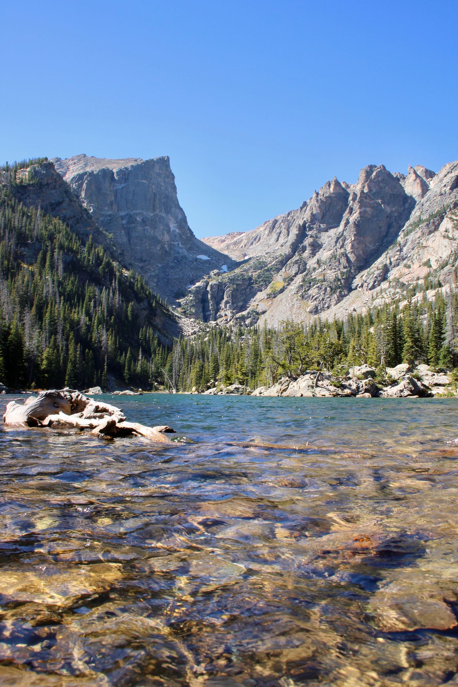 Crystal clear mountain lake waters reveal rocky bottom with majestic Rocky Mountain peaks rising in the background.