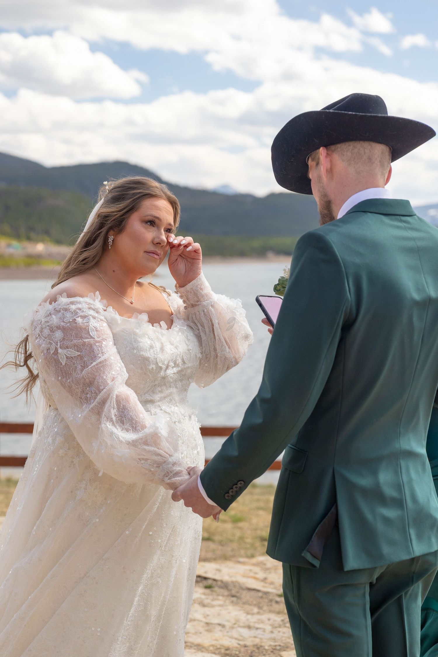 Bride wiping tears while exchanging vows with groom in green suit and cowboy hat at outdoor lakeside wedding ceremony.