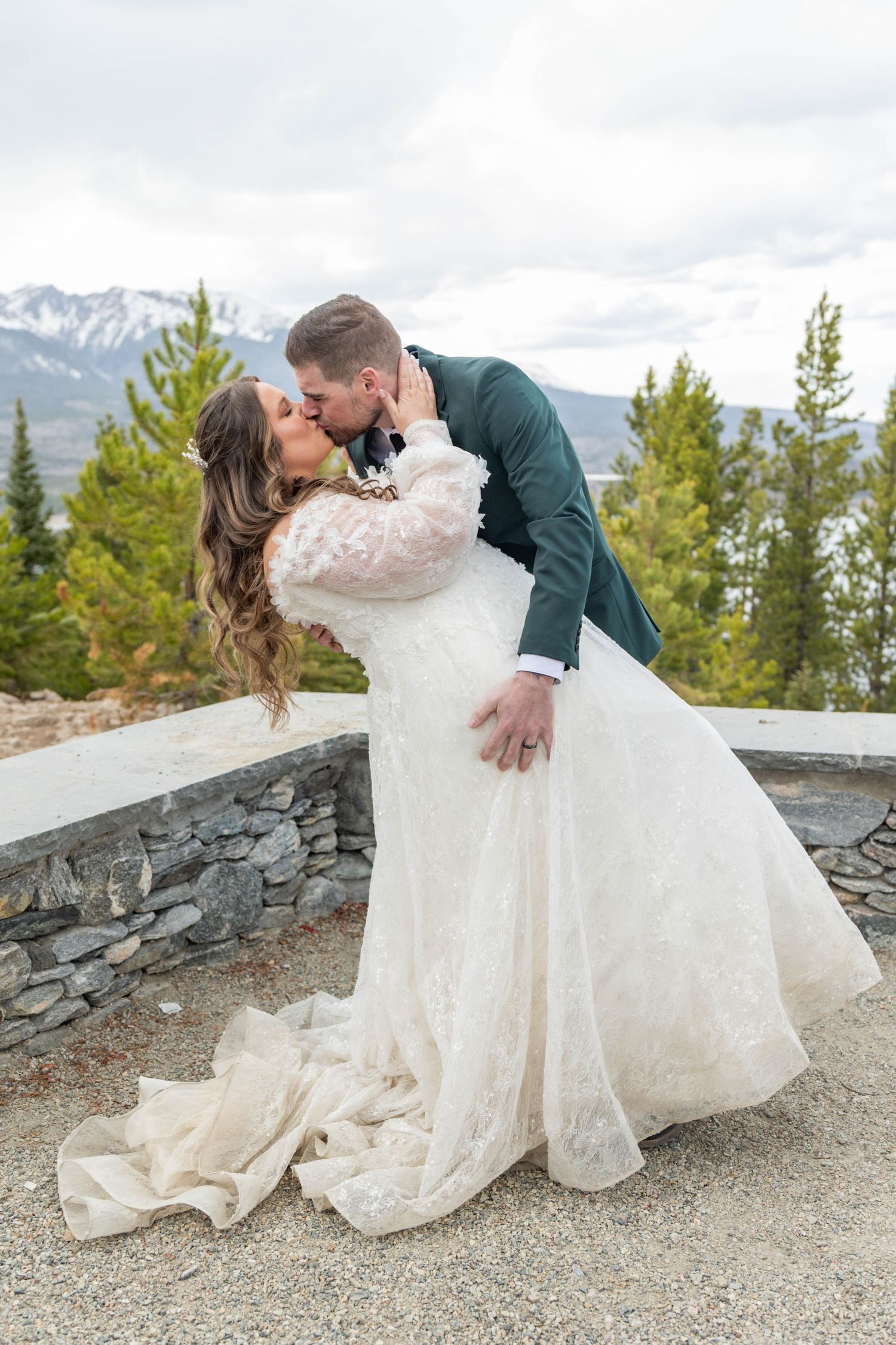 Bride and groom share a romantic dip kiss in lace wedding dress and teal suit with mountain backdrop.