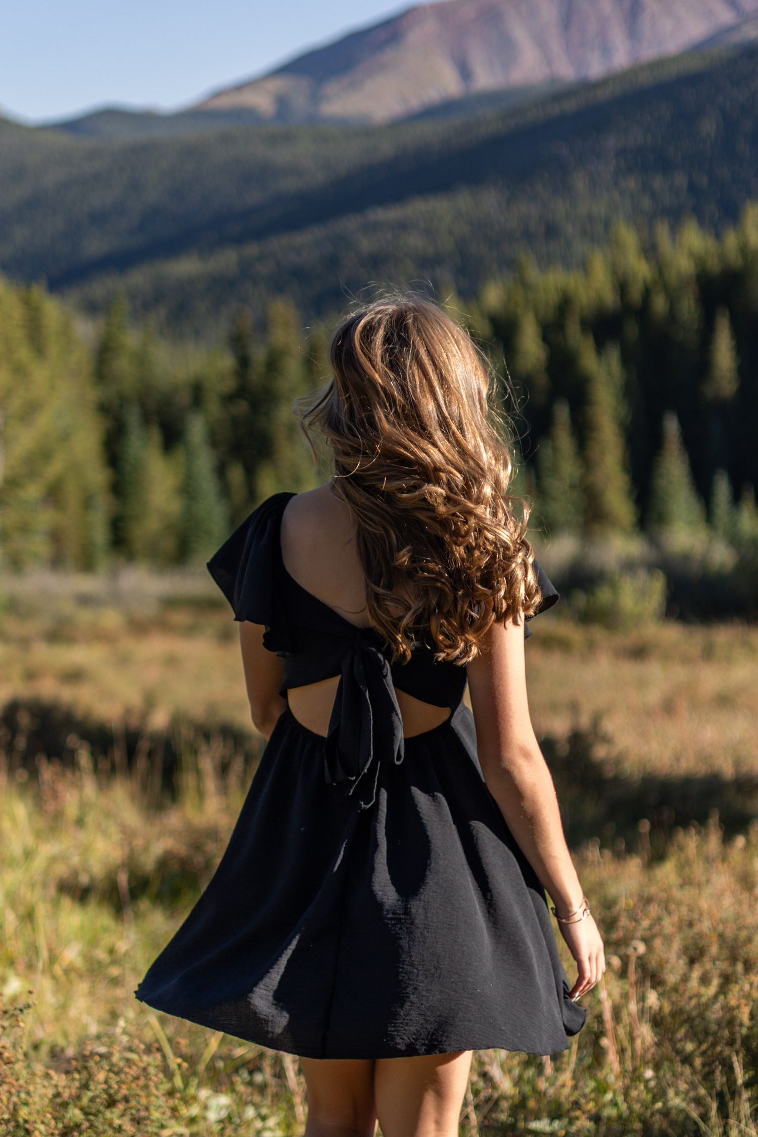 A person in a black dress twirls in a mountain meadow with evergreen forests and rugged peaks in the background.