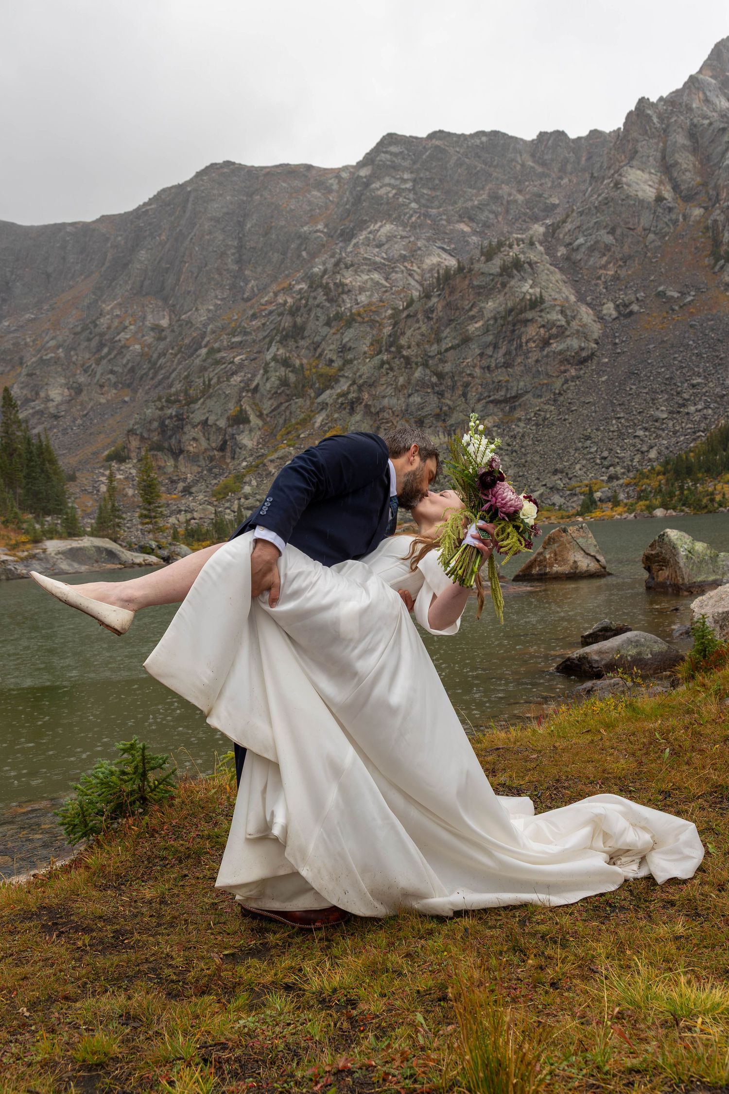 Groom dips bride in a white gown for a kiss beside a mountain lake with rocky peaks, holding a colorful bouquet during an elopement.