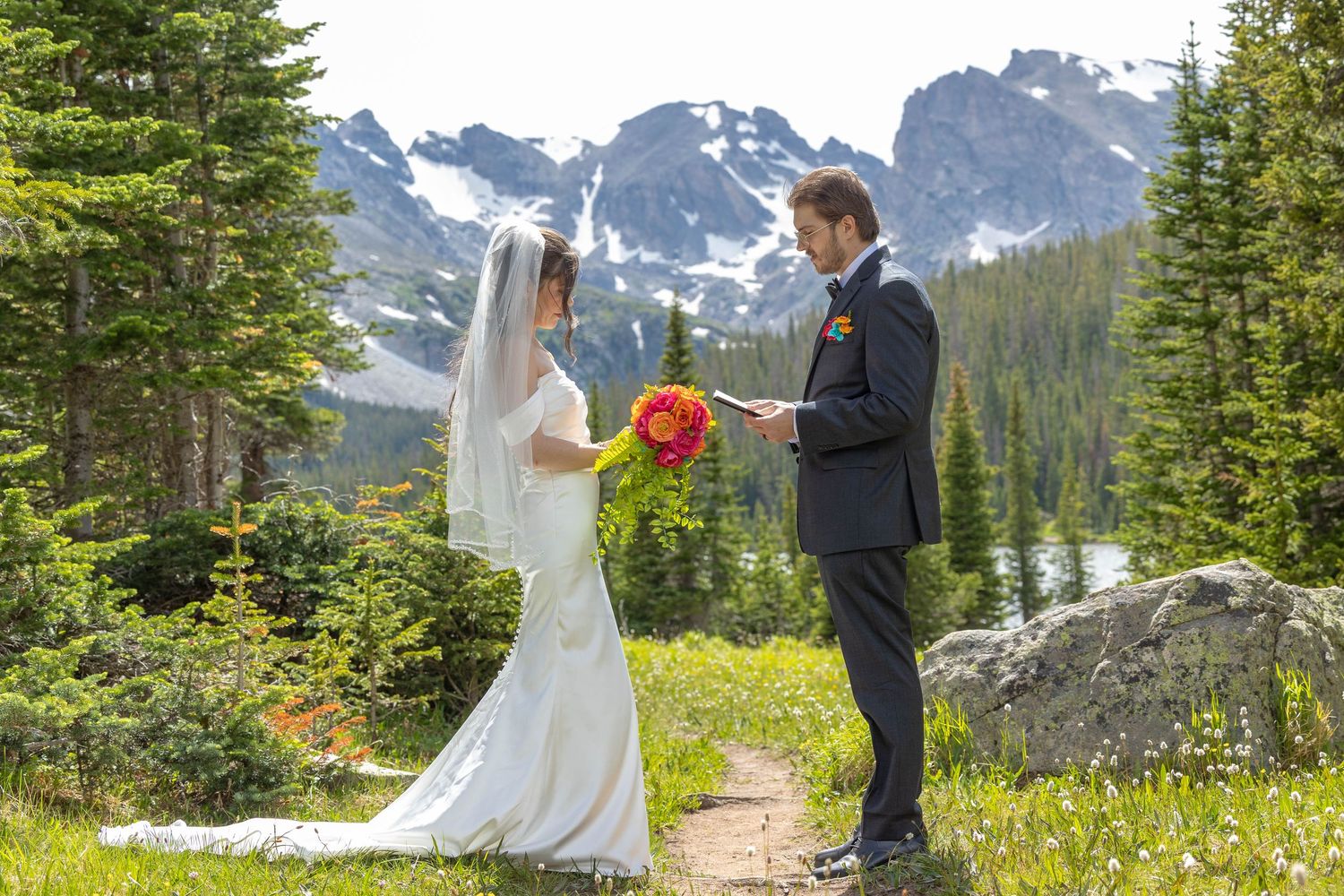 Bride and groom exchange vows in a mountain meadow wedding ceremony surrounded by evergreen trees and snow-capped peaks.